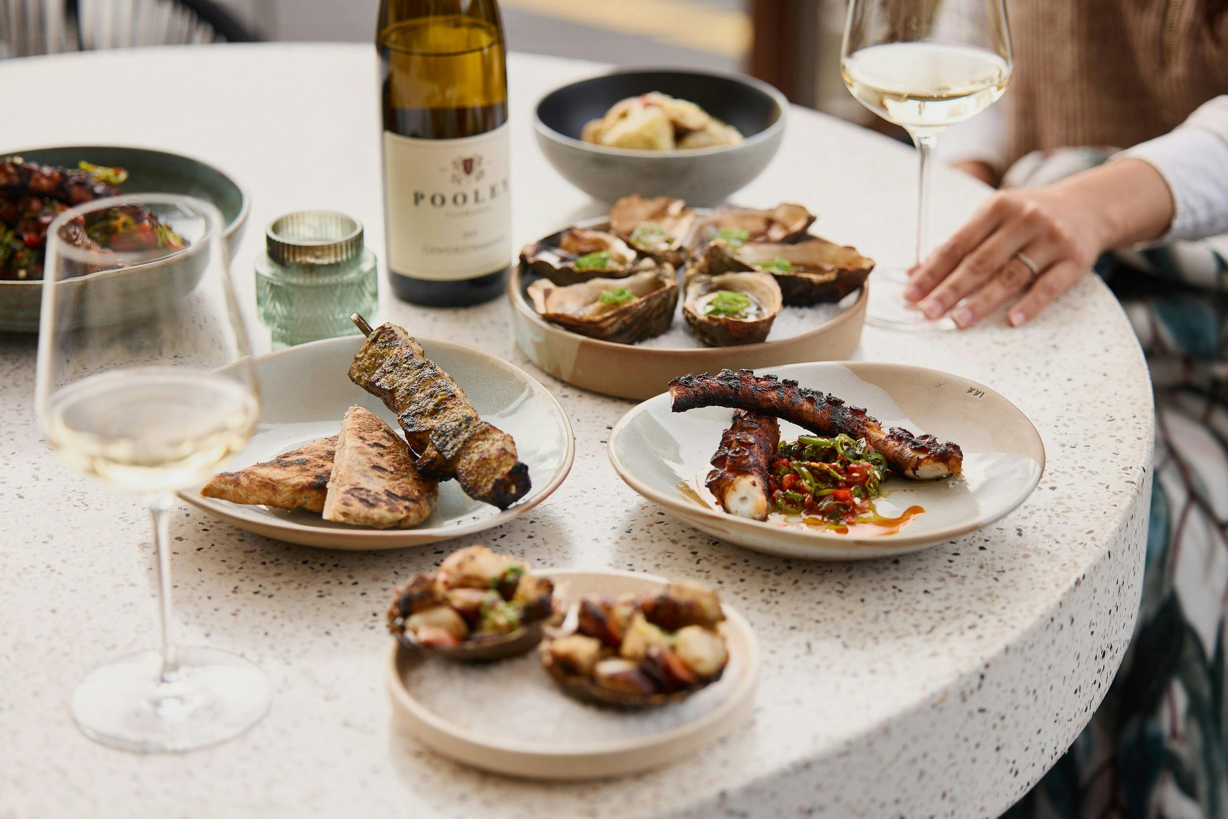 A selection of seafood dishes sit on a white table, accompanied by a Pooley Wine bottle and glass.