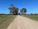 Dull brown gravel road, bushy grass, fences, paddocks, gum trees, blue sky, mountains in background