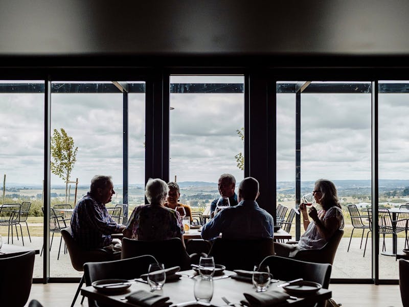 Guests sitting at a table by the ceiling to floor windows. They are chatting and enjoying wine.