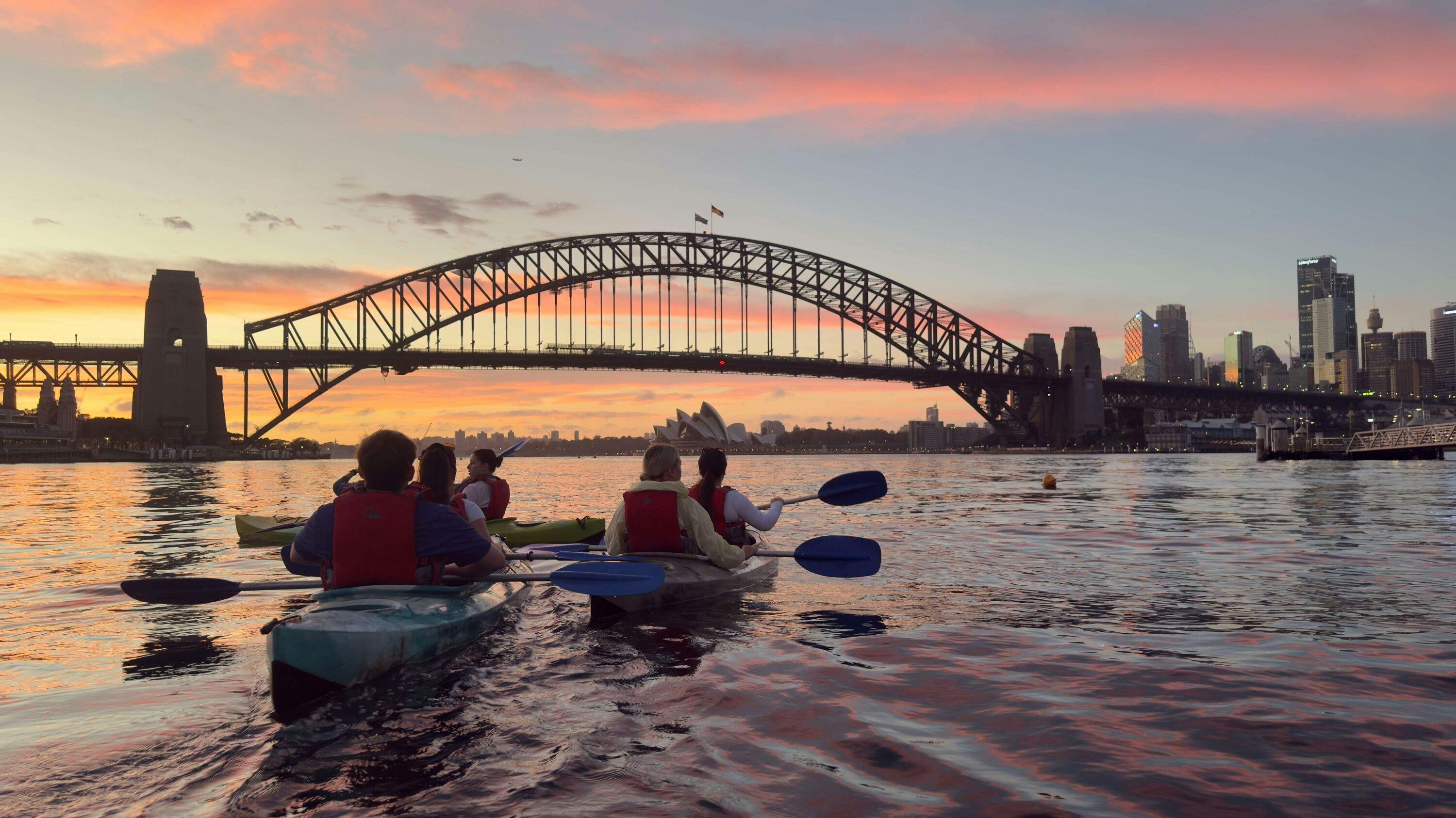 雙人皮划艇駛近雪梨港灣大橋，日出時分，雪梨歌劇院清晰可見。