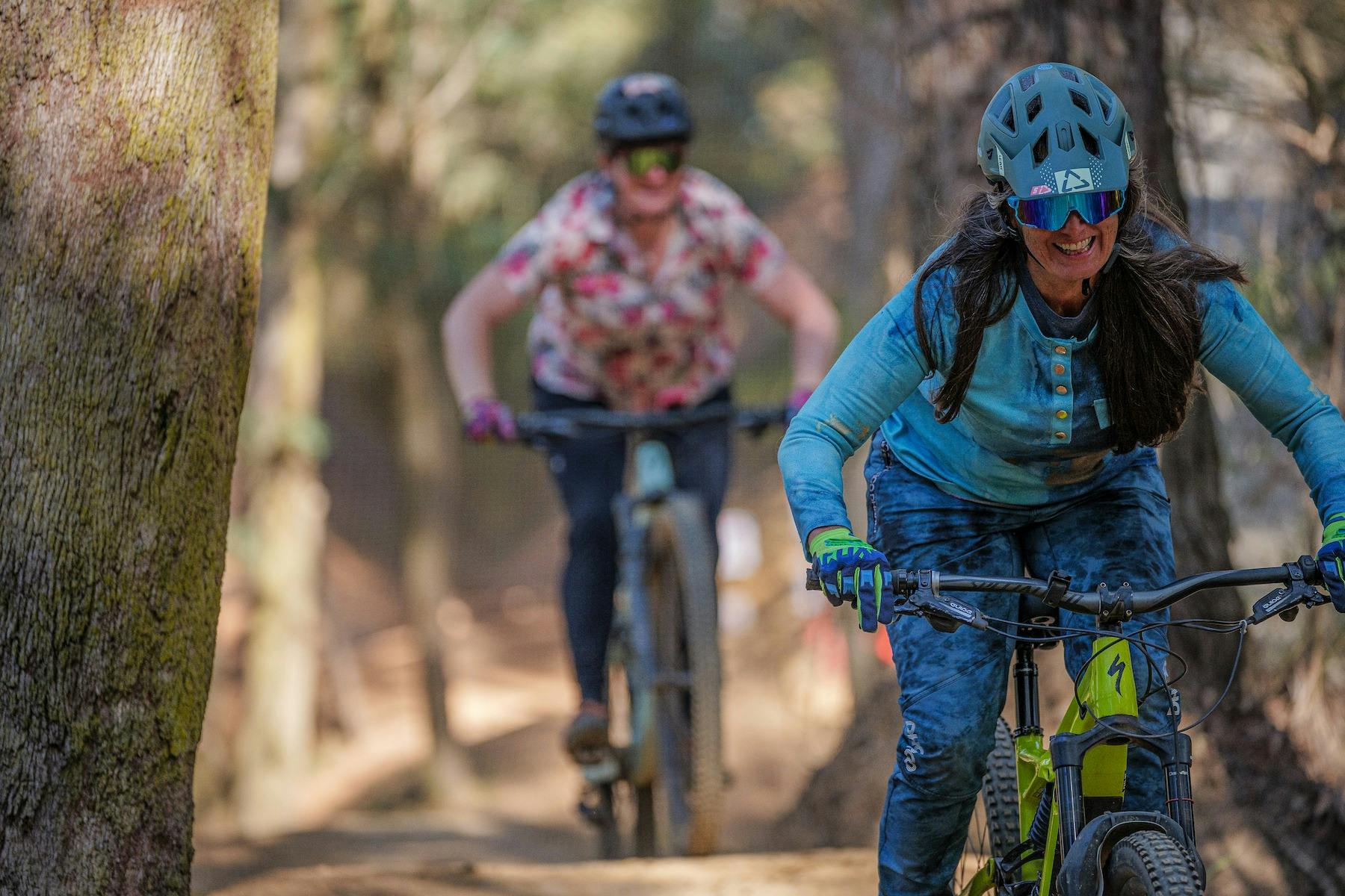 A female rider wearing blue is followed down a mountain bike trail by another female rider.