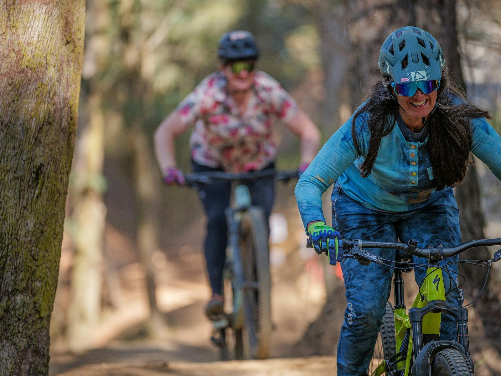 A female rider wearing blue is followed down a mountain bike trail by another female rider.