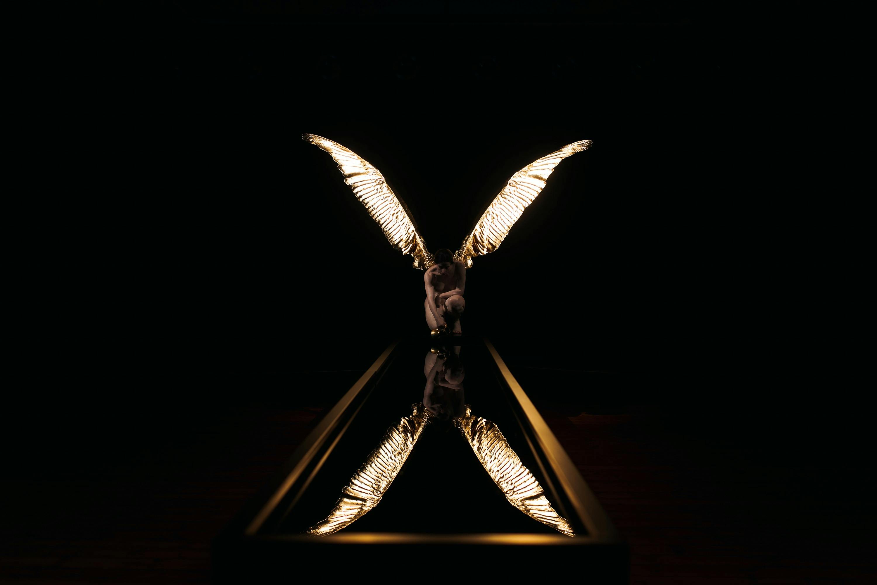 A sculpture of a kneeling Greek goddess with gold wings, reflected in a pool of water.