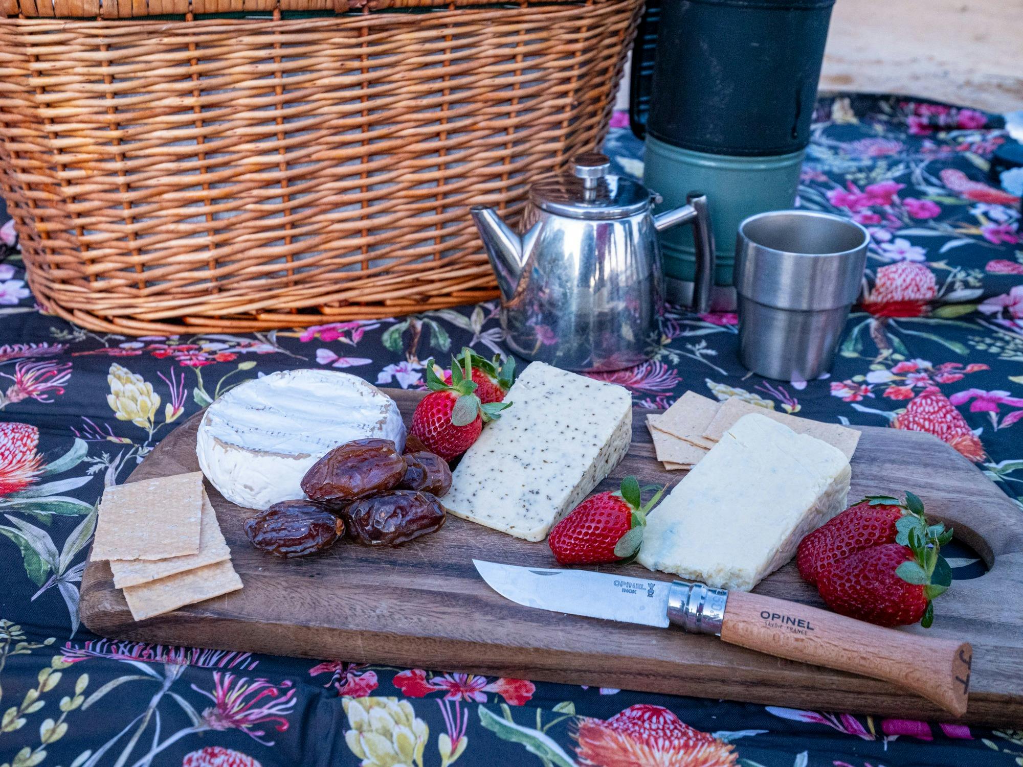 A picnic rug with a teapot, cups, thermos, cheese board, and picnic basket.