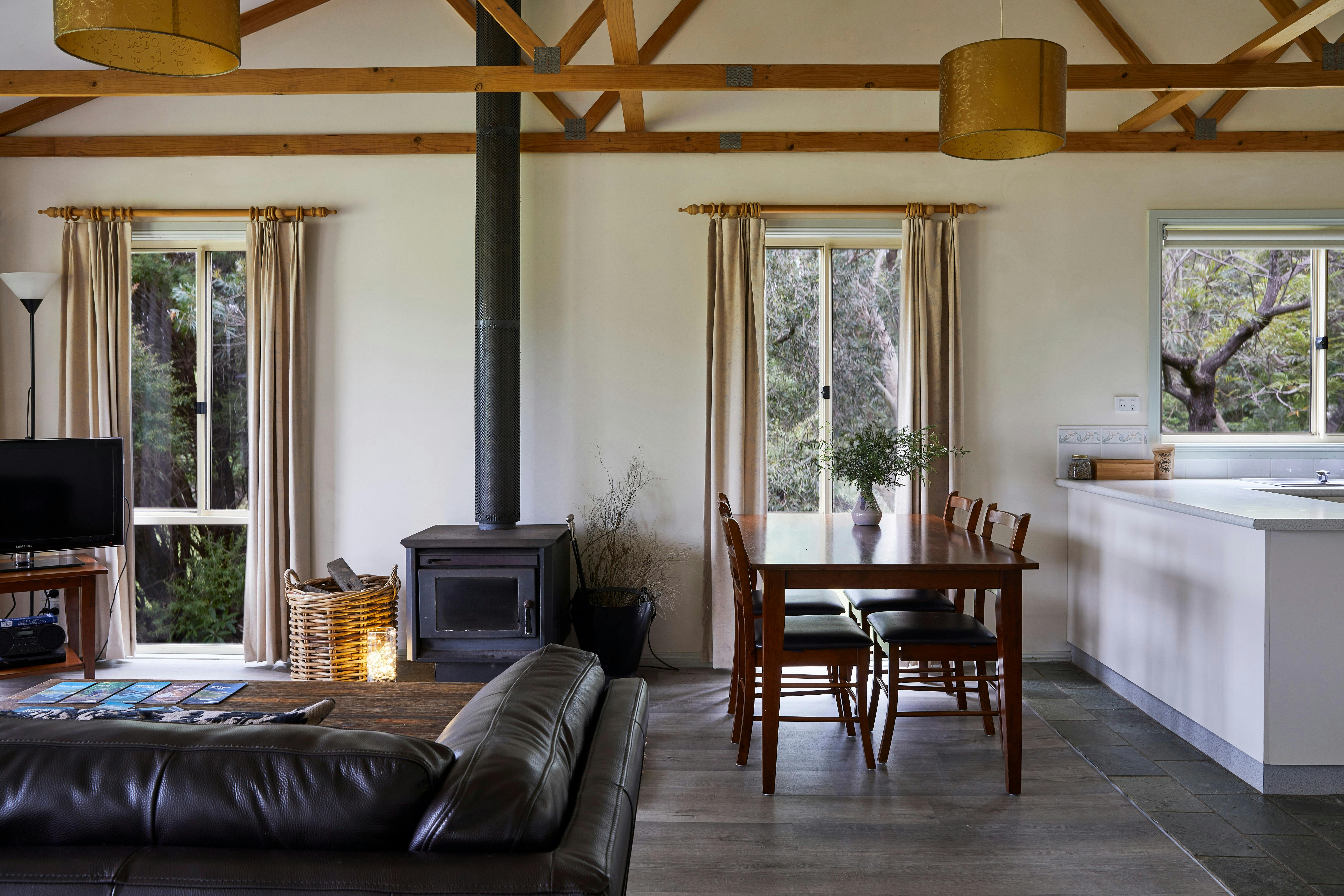Cottage interior shows wood burner, sofa, dining table and edge of kitchen surface. Natural  light