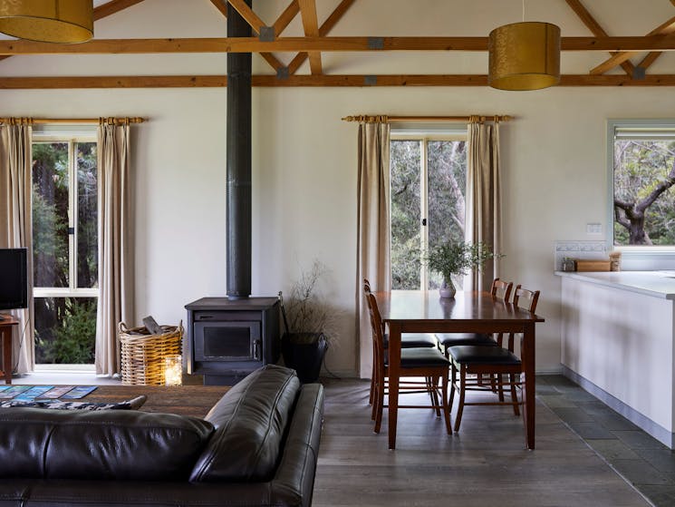 Cottage interior shows wood burner, sofa, dining table and edge of kitchen surface. Natural  light