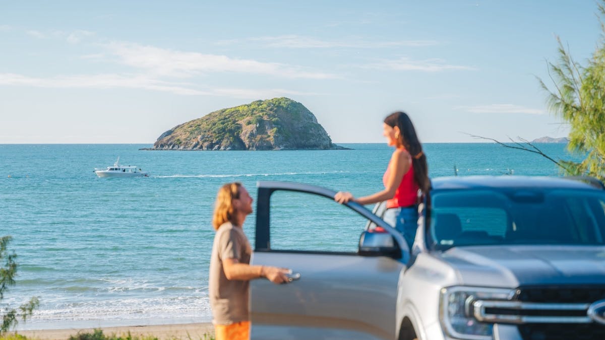 Two tourists pause to look at a boat and a distant mountain rising from the sea.