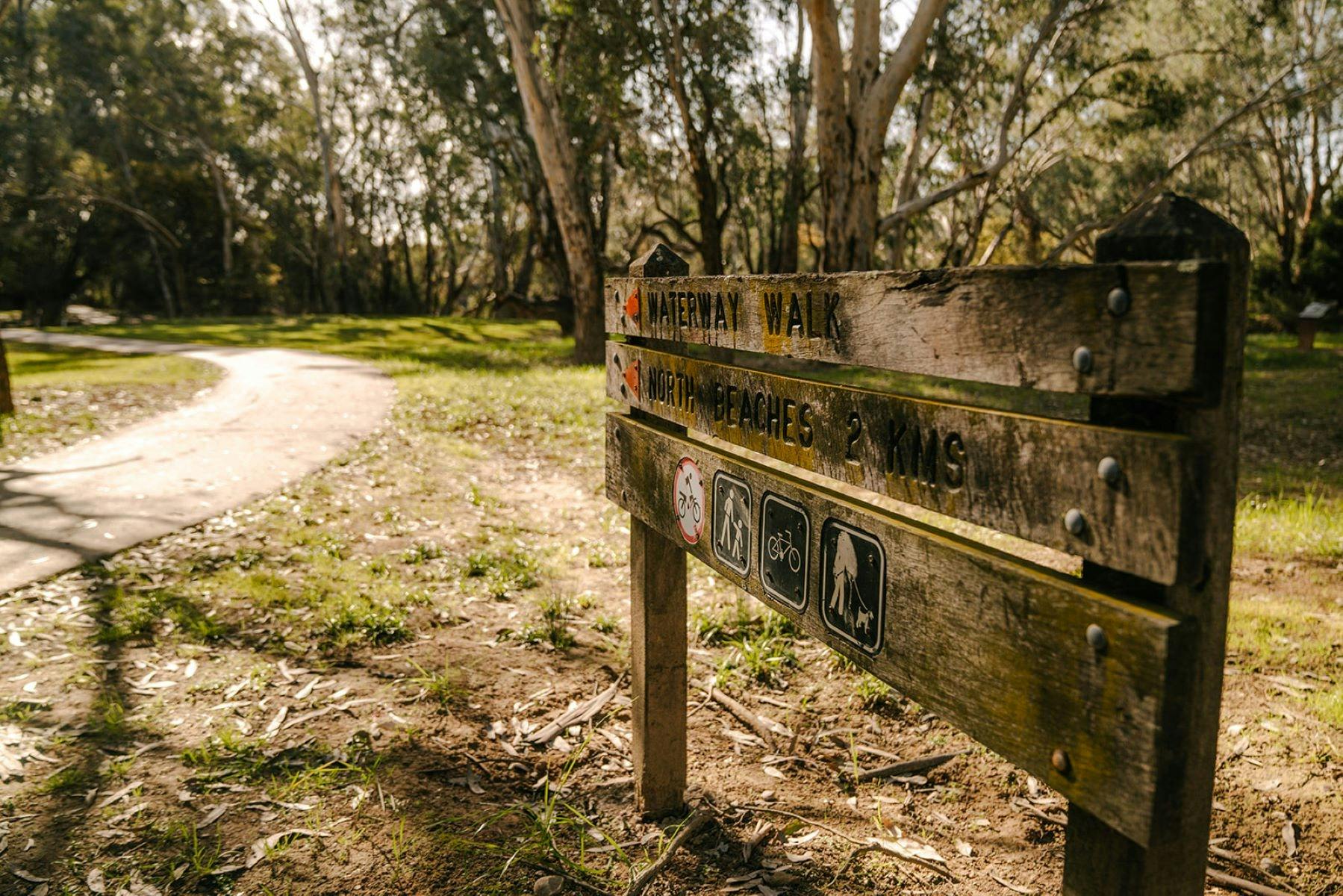Brown sign with directions for river walk trails on the edge of a bike path winding through grass