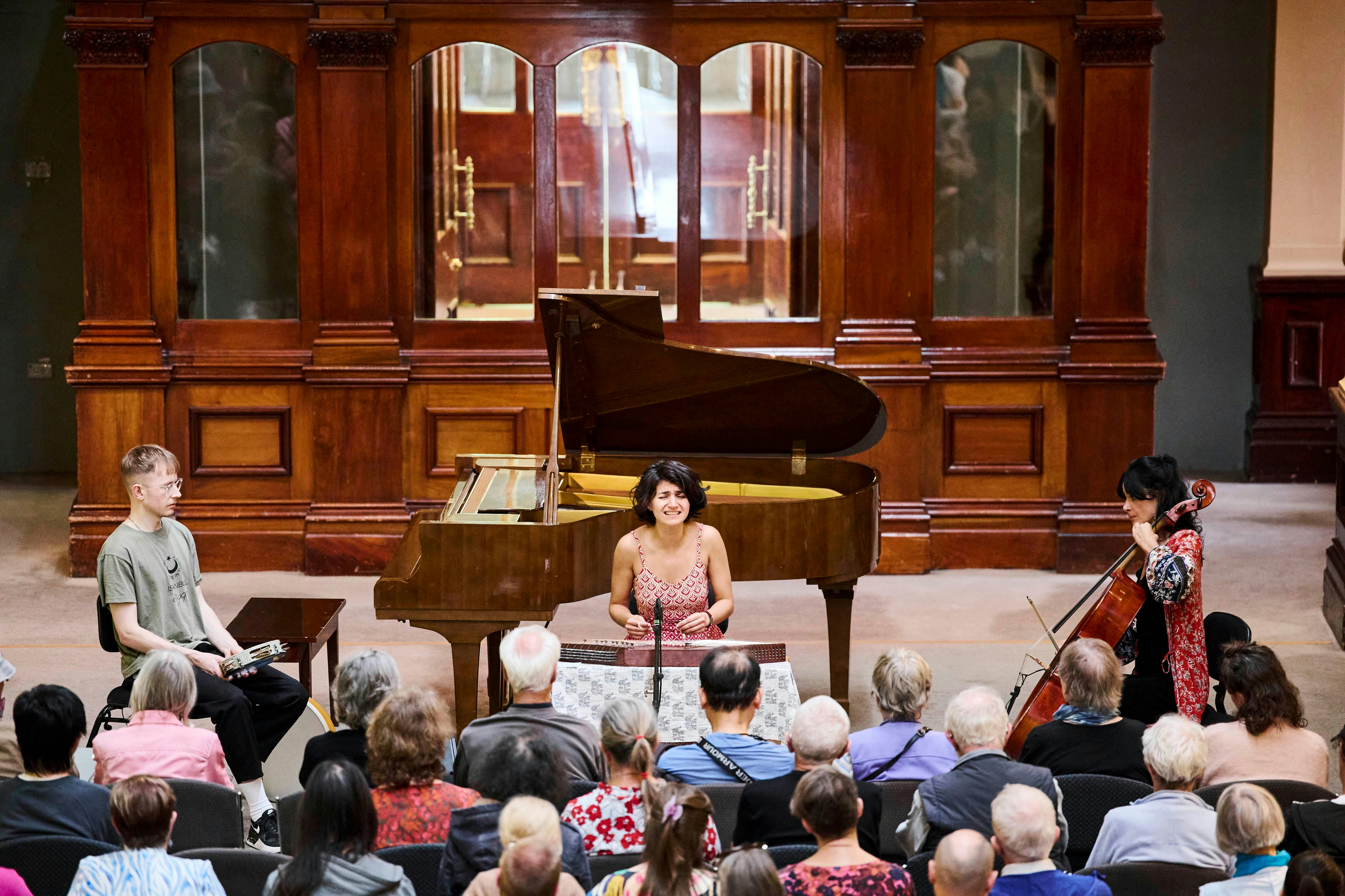 An audience watches a trio of musicians perform in a museum, with collections displayed behind.