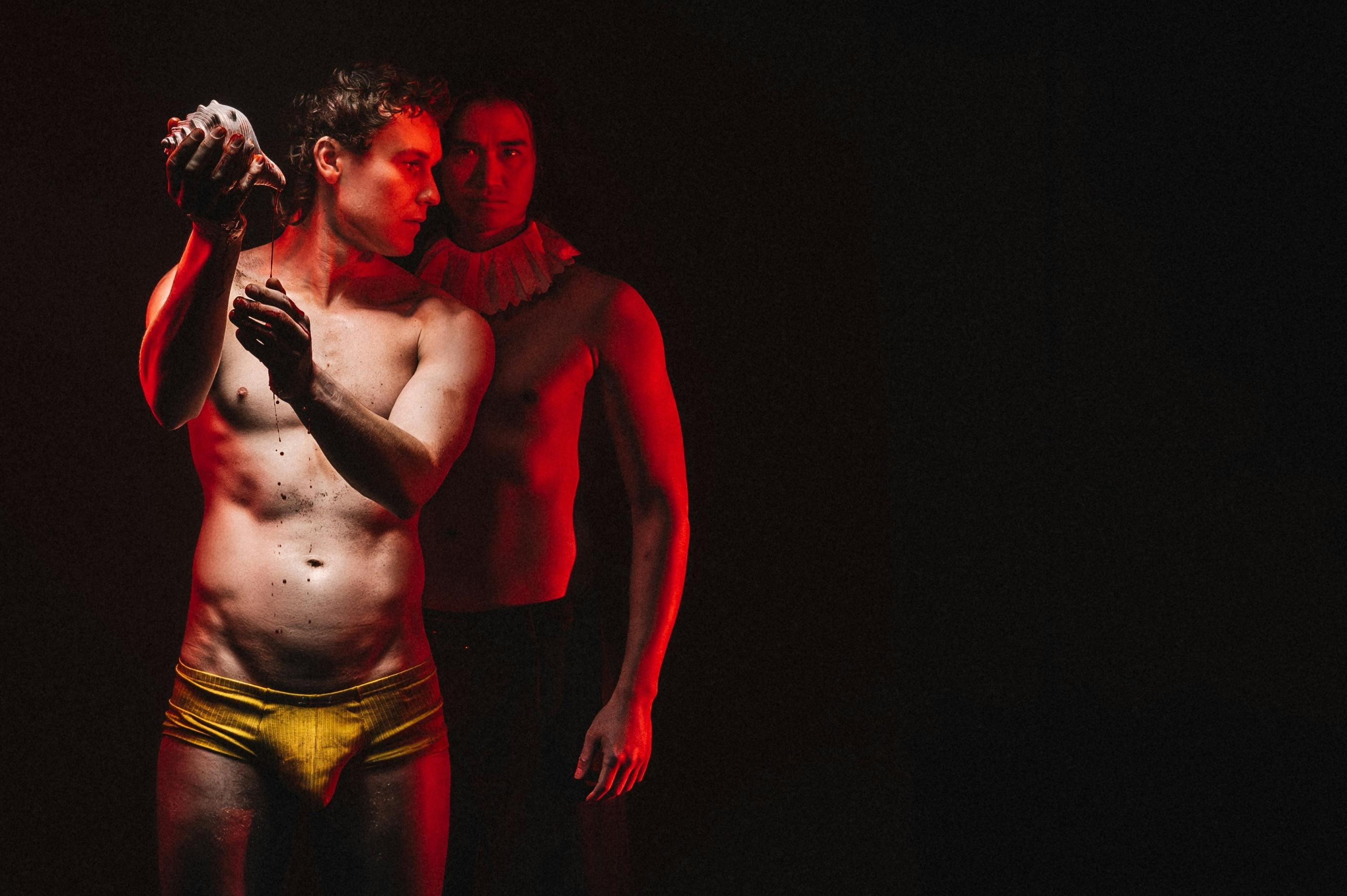 Two topless men stand in fron of eachother, one of them pours sea water from a conch shell.