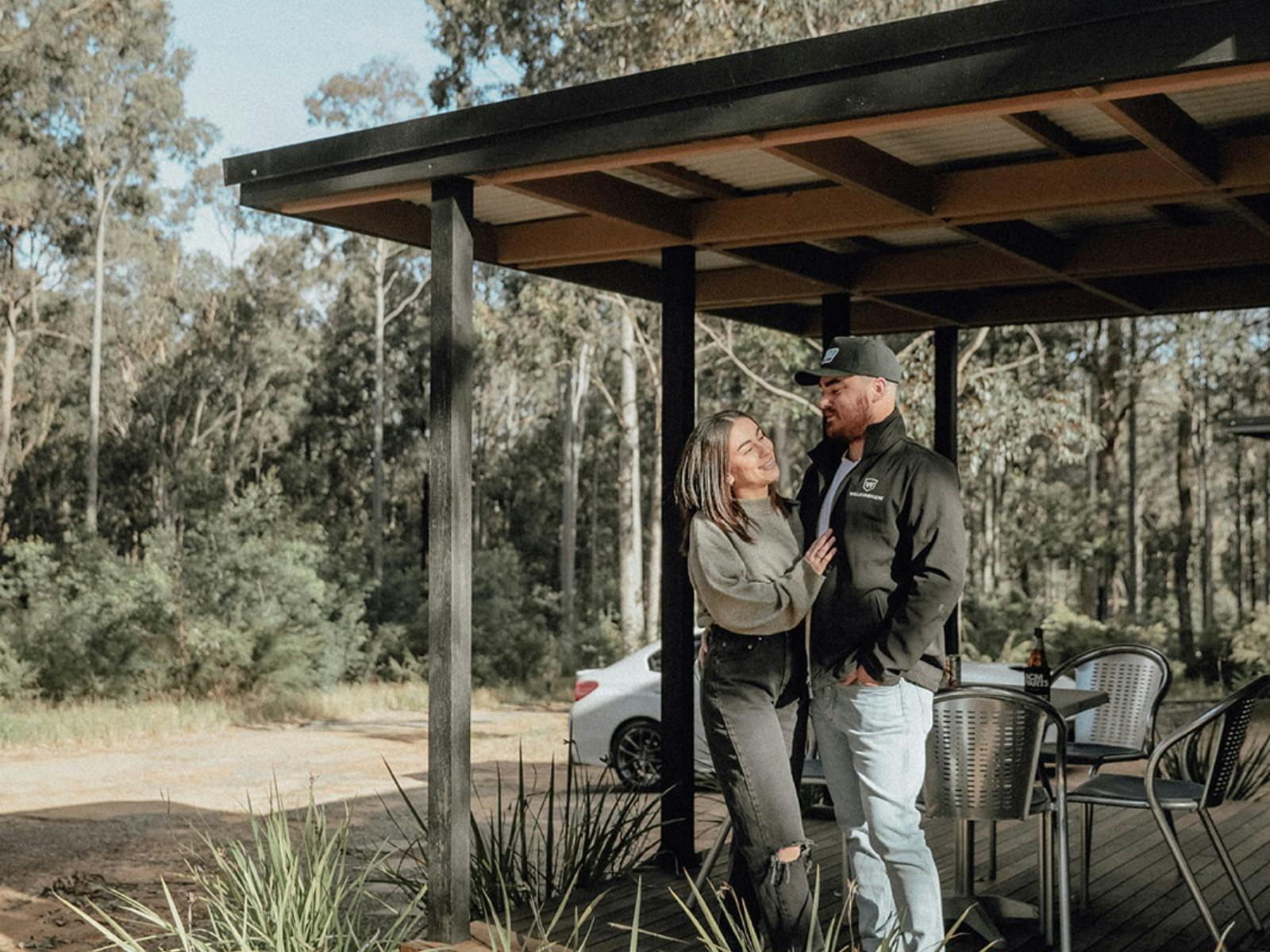 호주 숲으로 둘러싸인 헌터 밸리 리트리트 아파트의 파티오에서 남자와 여자가 껴안고 있습니다.