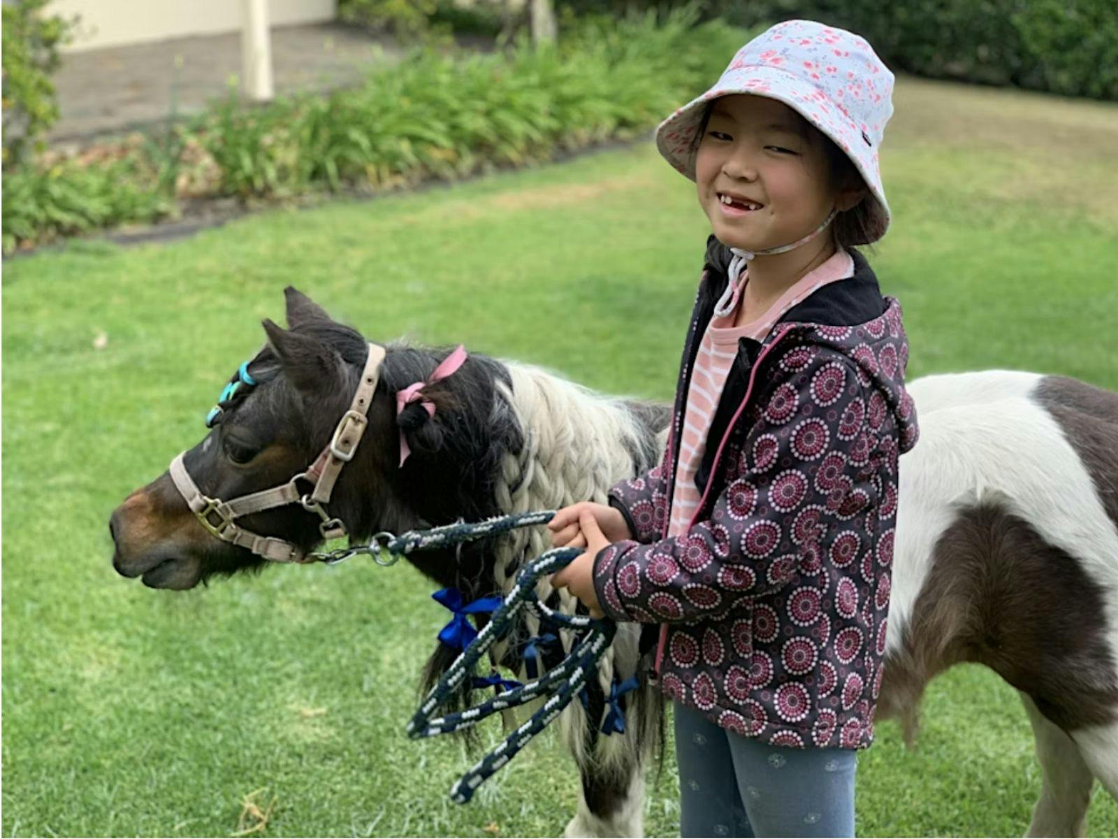 Young girl smiling brightly while walking next to a miniature horse in an outdoor farm setting