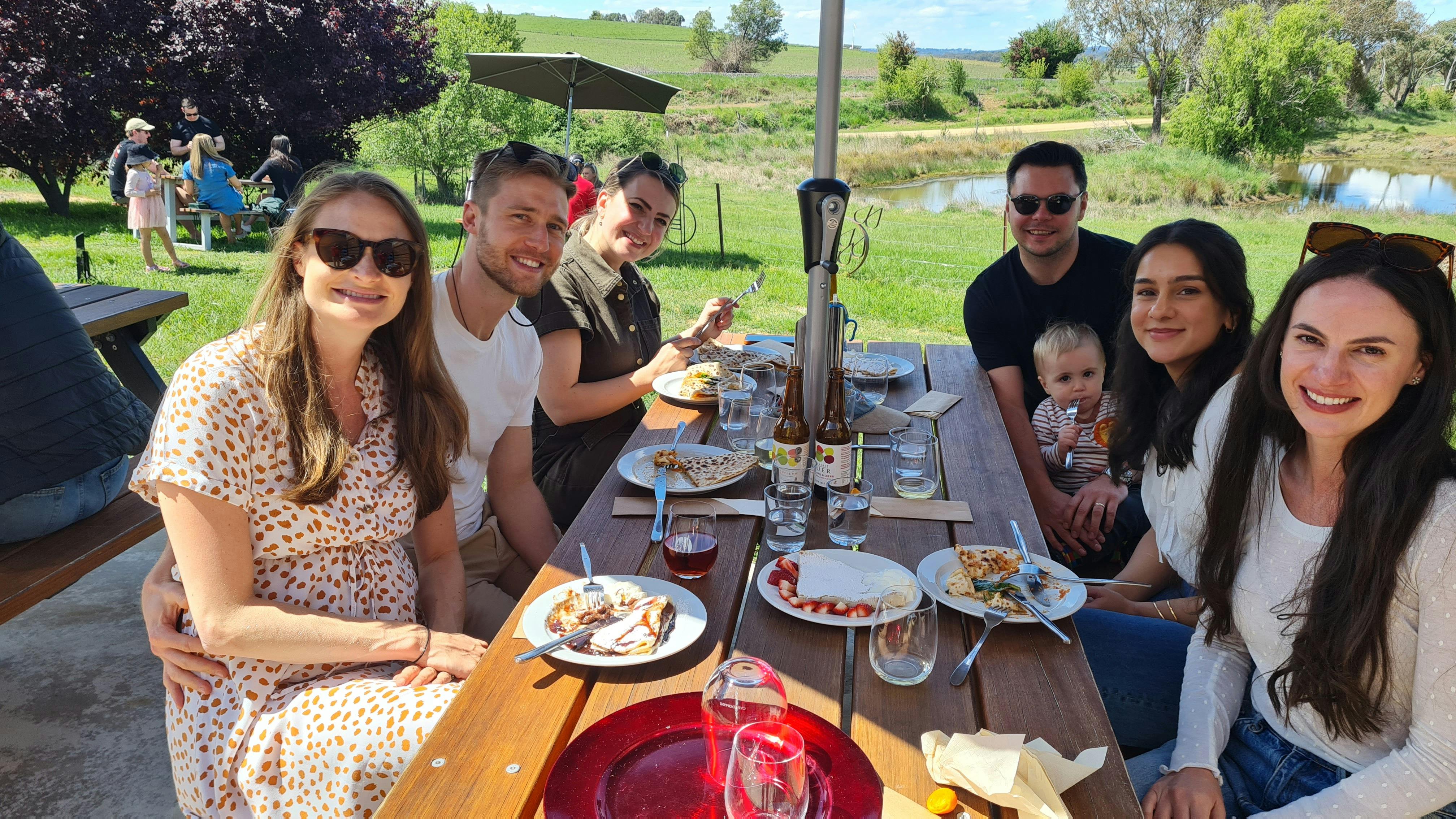 Smiling people sit around a table with crepes and cider on the table. Lush green grass and a dam.