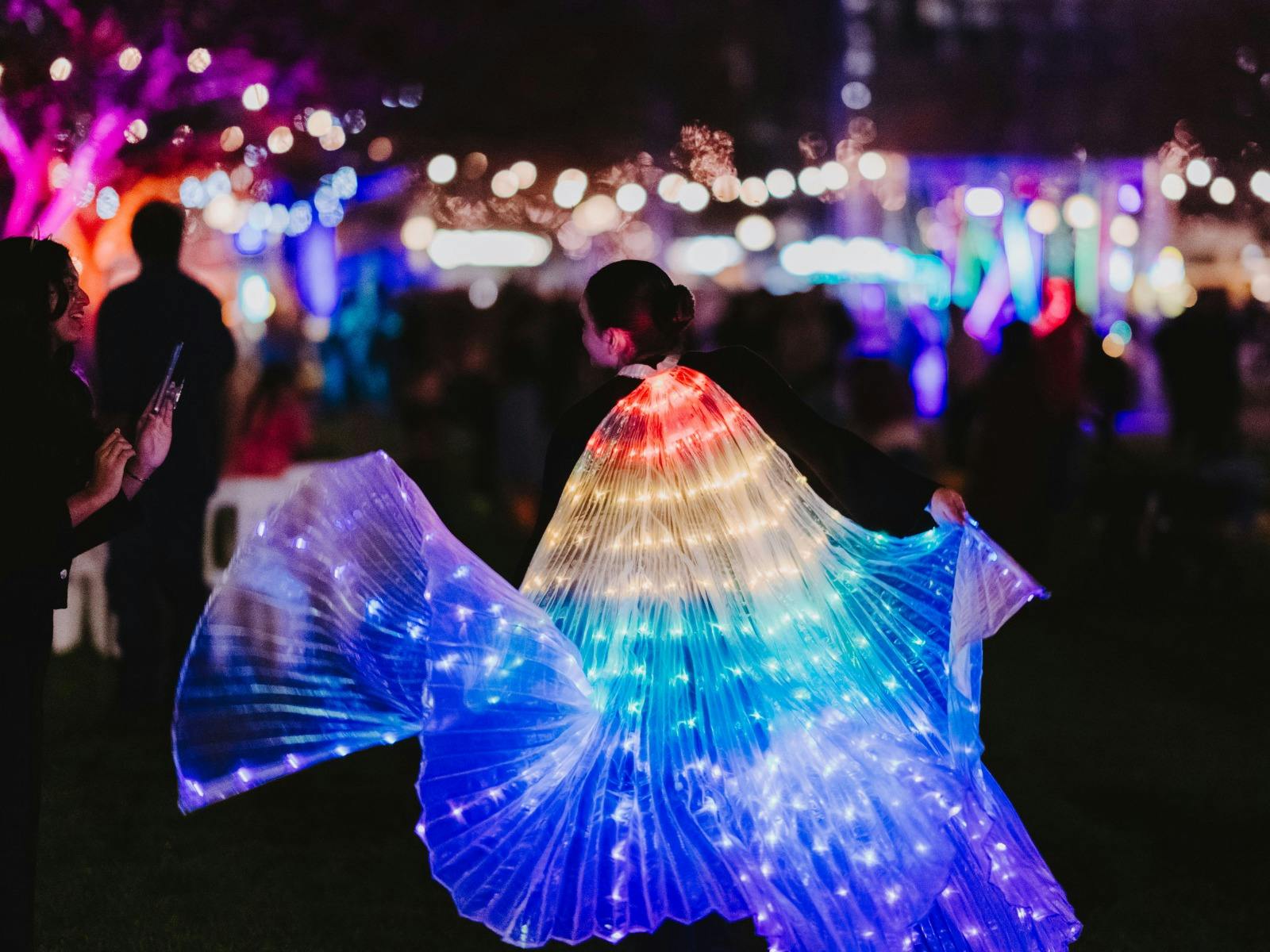 Performer spinning with illuminated, multicolored wing costume in a lit festival crowd.