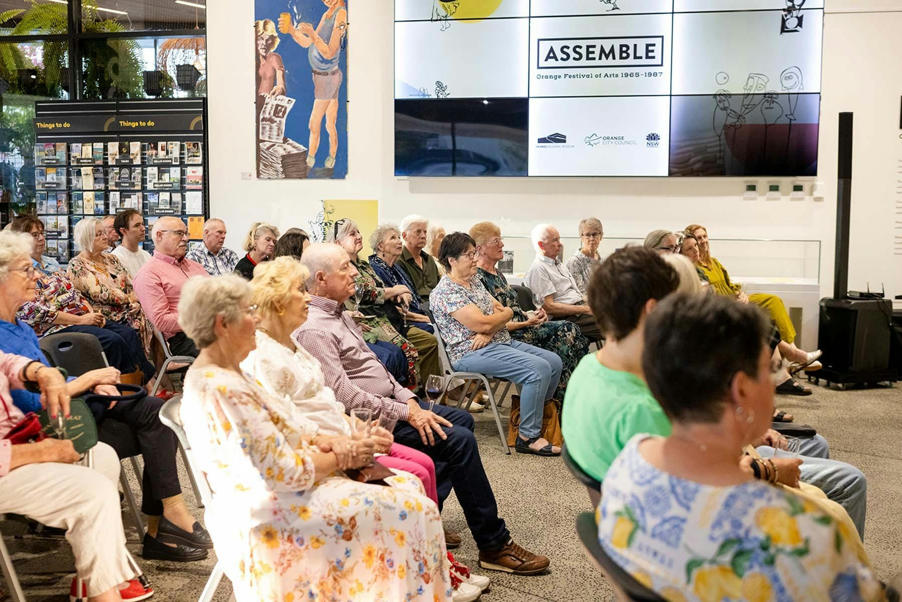 People sit in a small crowd listening to a speaker at an exhibition opening