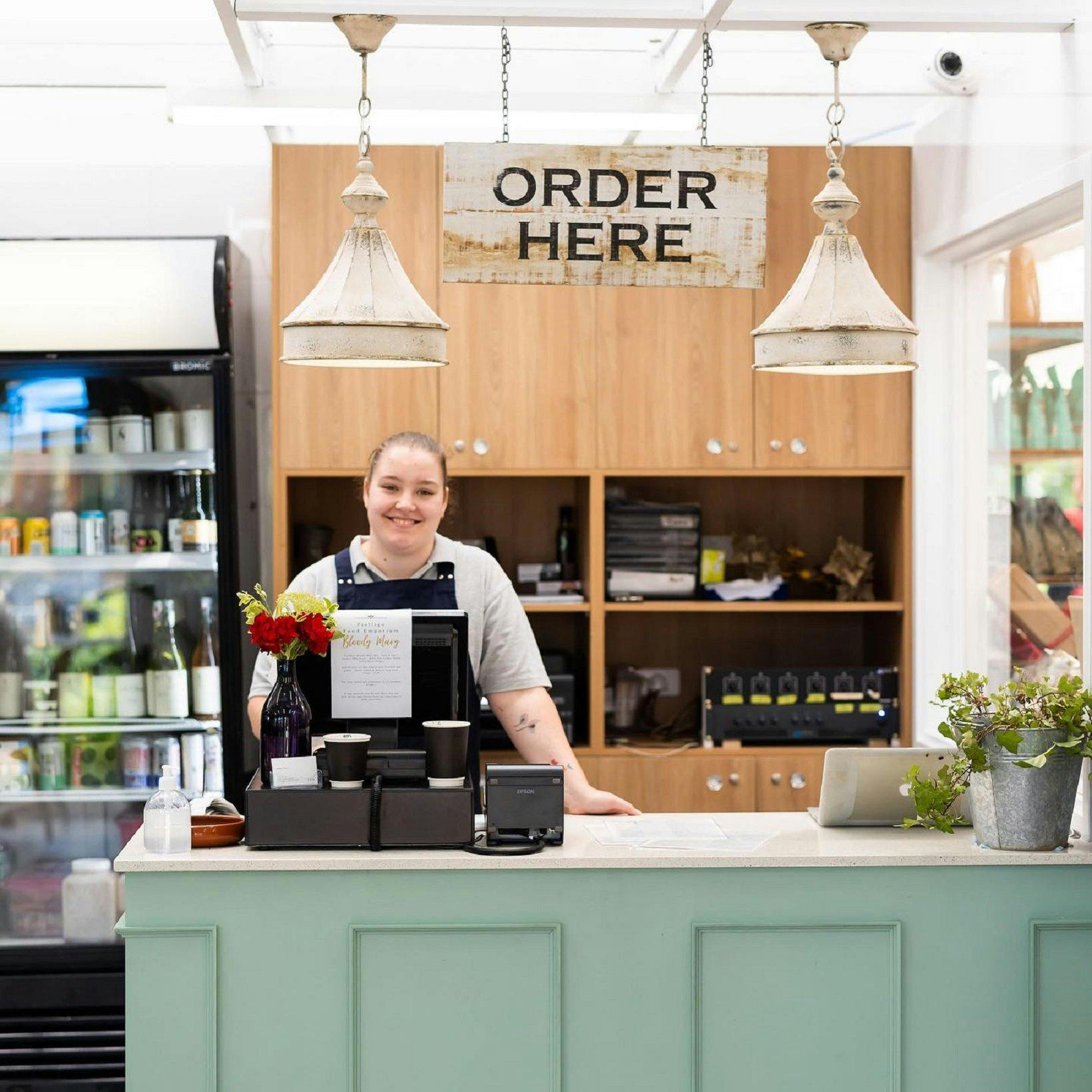 A woman behind a cafe counter underneath a sign that says 'order here',  ready to serve customers