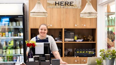 A woman behind a cafe counter underneath a sign that says 'order here', ready to serve customers
