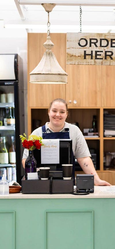 A woman behind a cafe counter underneath a sign that says 'order here', ready to serve customers