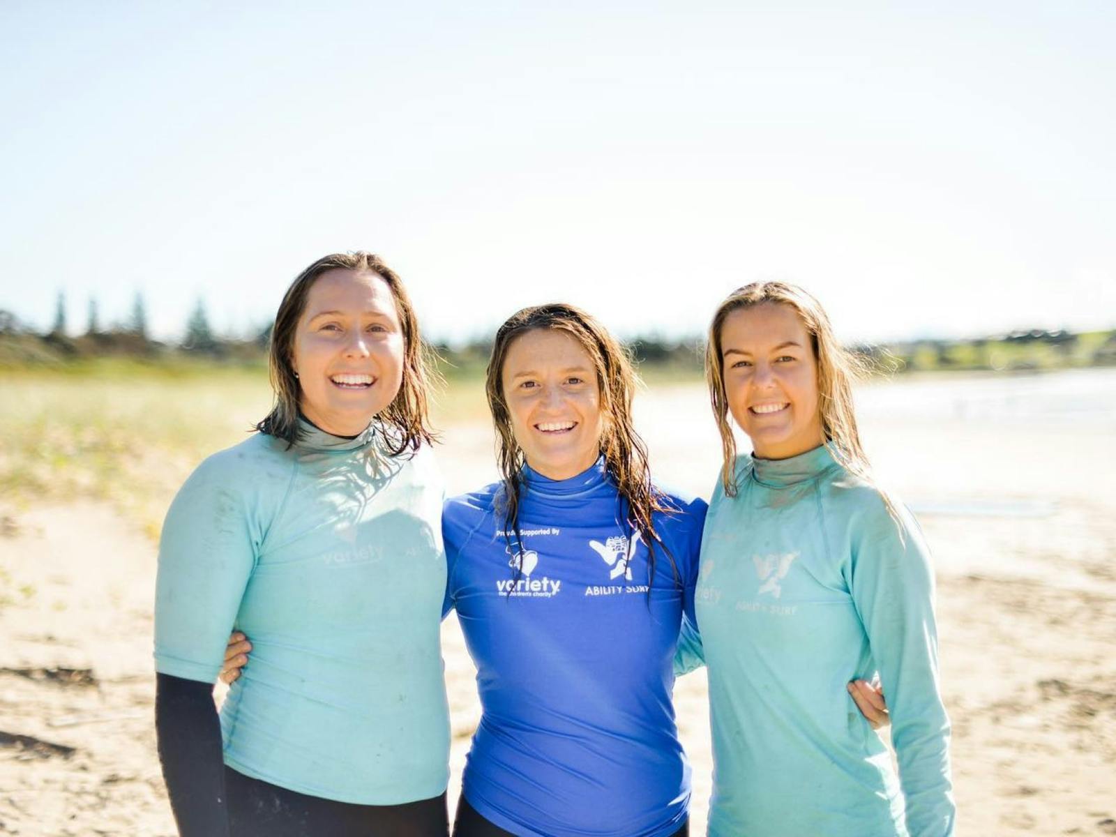 three women standing on beach in blue rash shirts smiling
