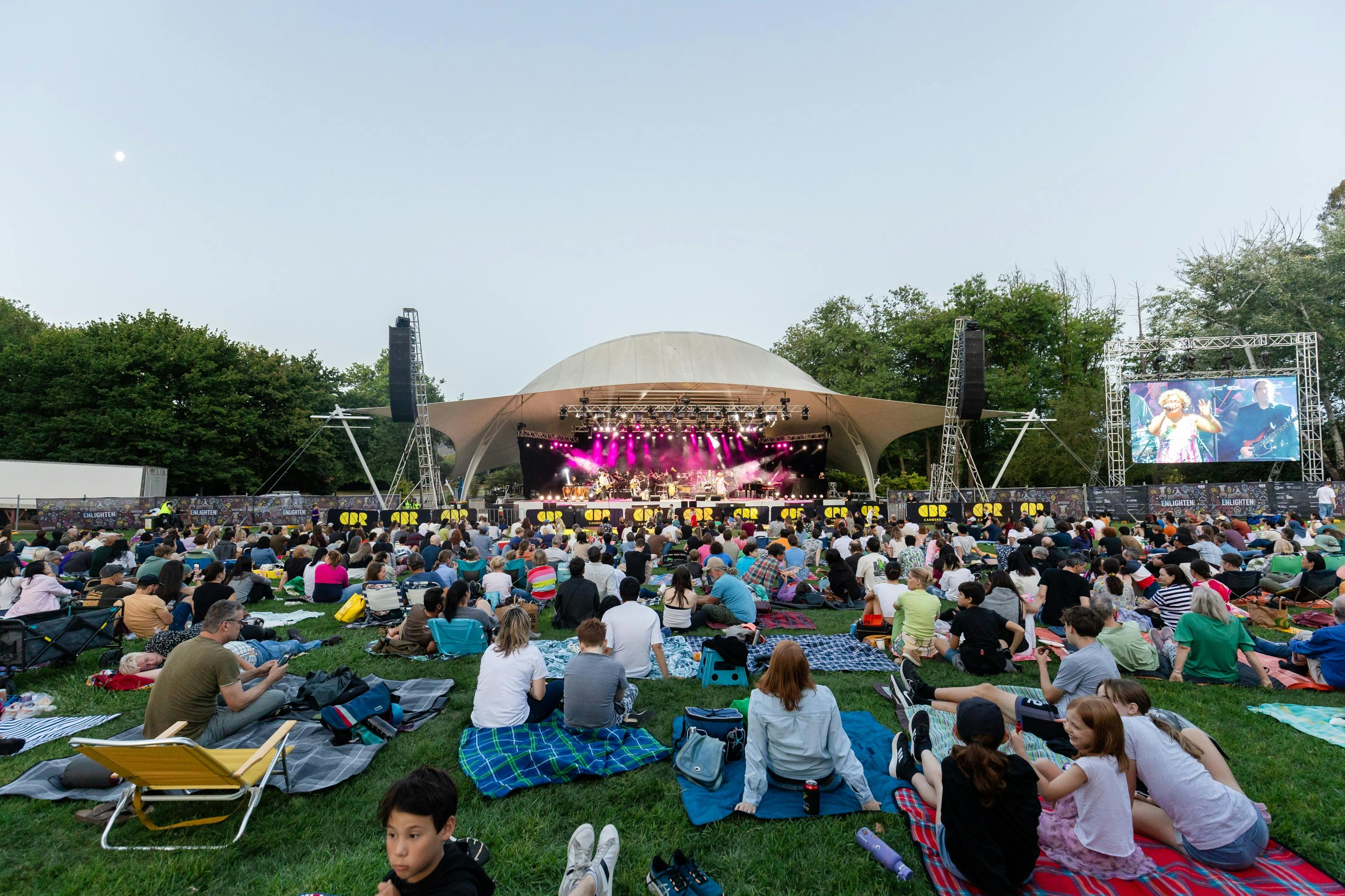 People sitting on grass, looking up at stage as they watch outdoor performance