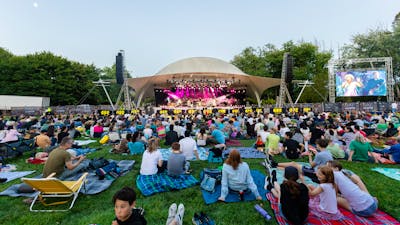People sitting on grass, looking up at stage as they watch outdoor performance