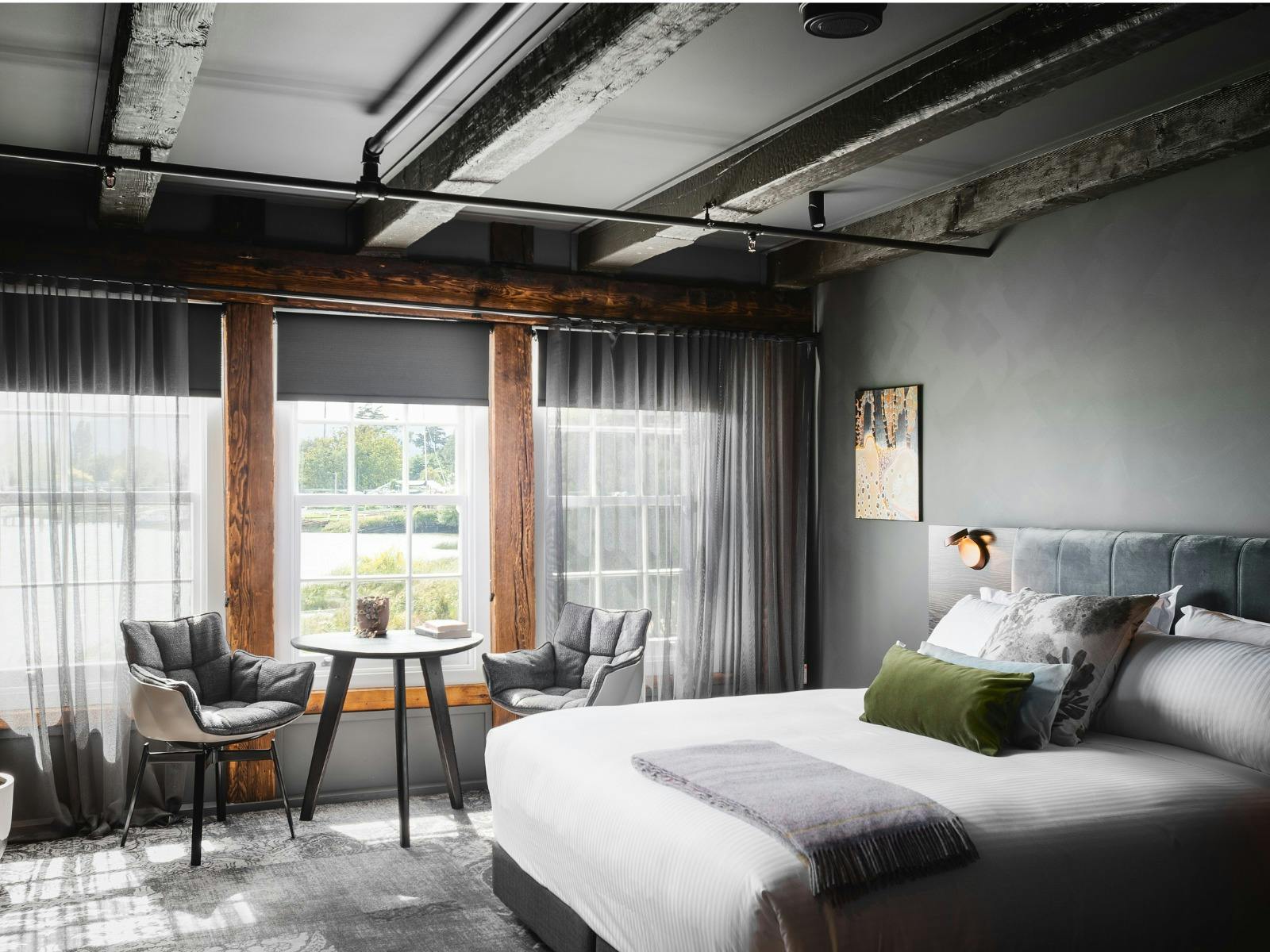 A king size bed, in a room with soft curtains and a table setting by the window