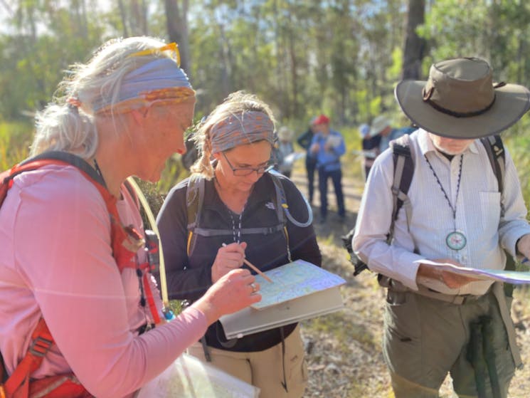 3 adults stand in the Aussie bush with maps in hand. 1 person is pointing out something on the map.