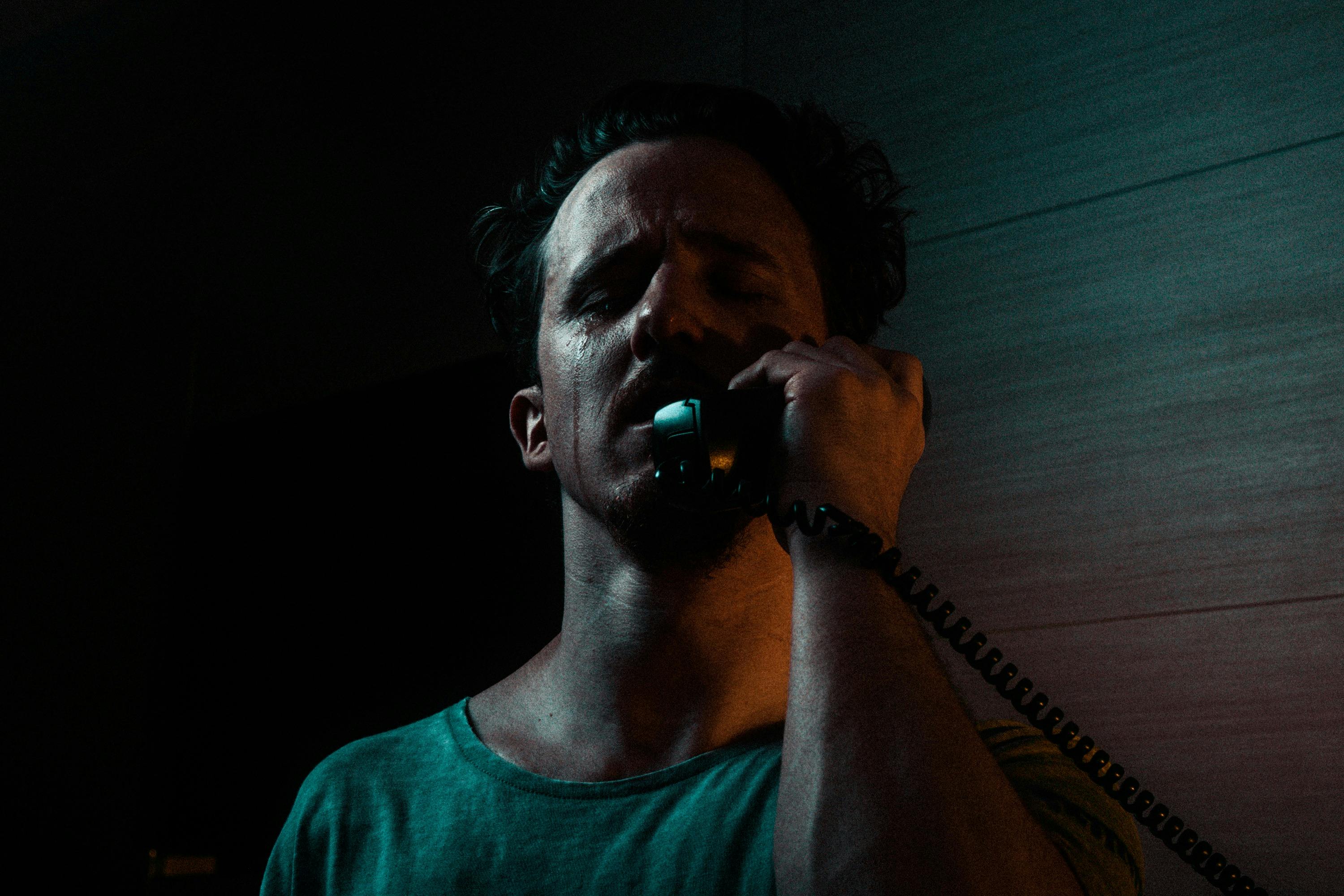 A man in a dimly lit room is holding a phone to his ear, looking distressed with tears on his face.
