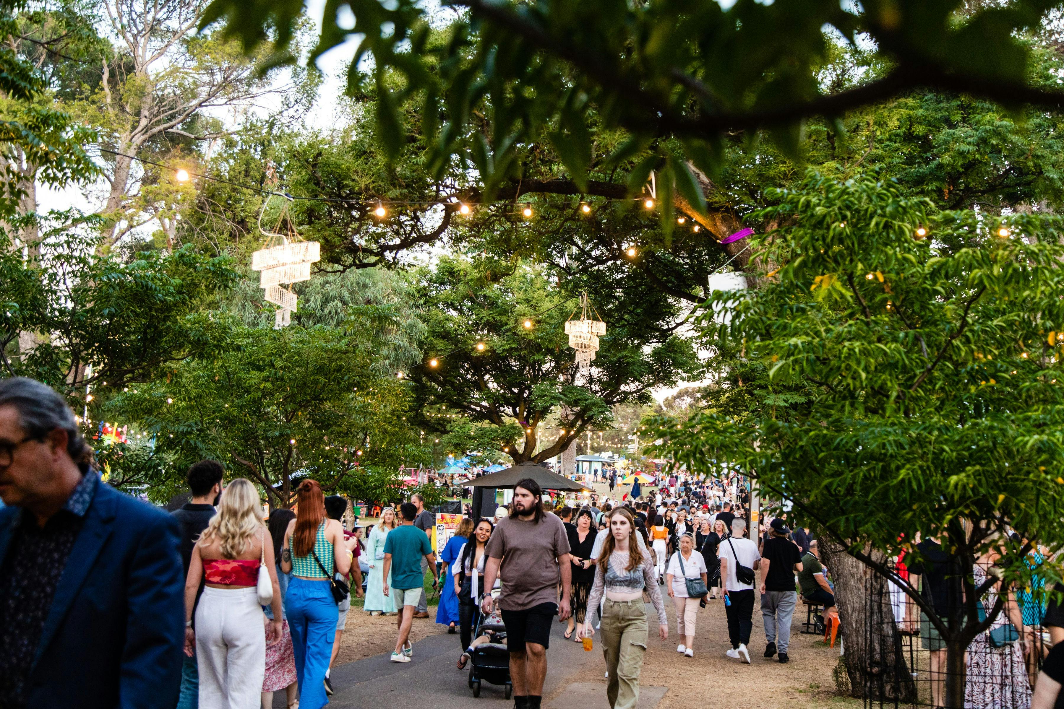 Patrons walking on concrete path through Gluttony hub. Overhead is lots of greenery and warm lights.