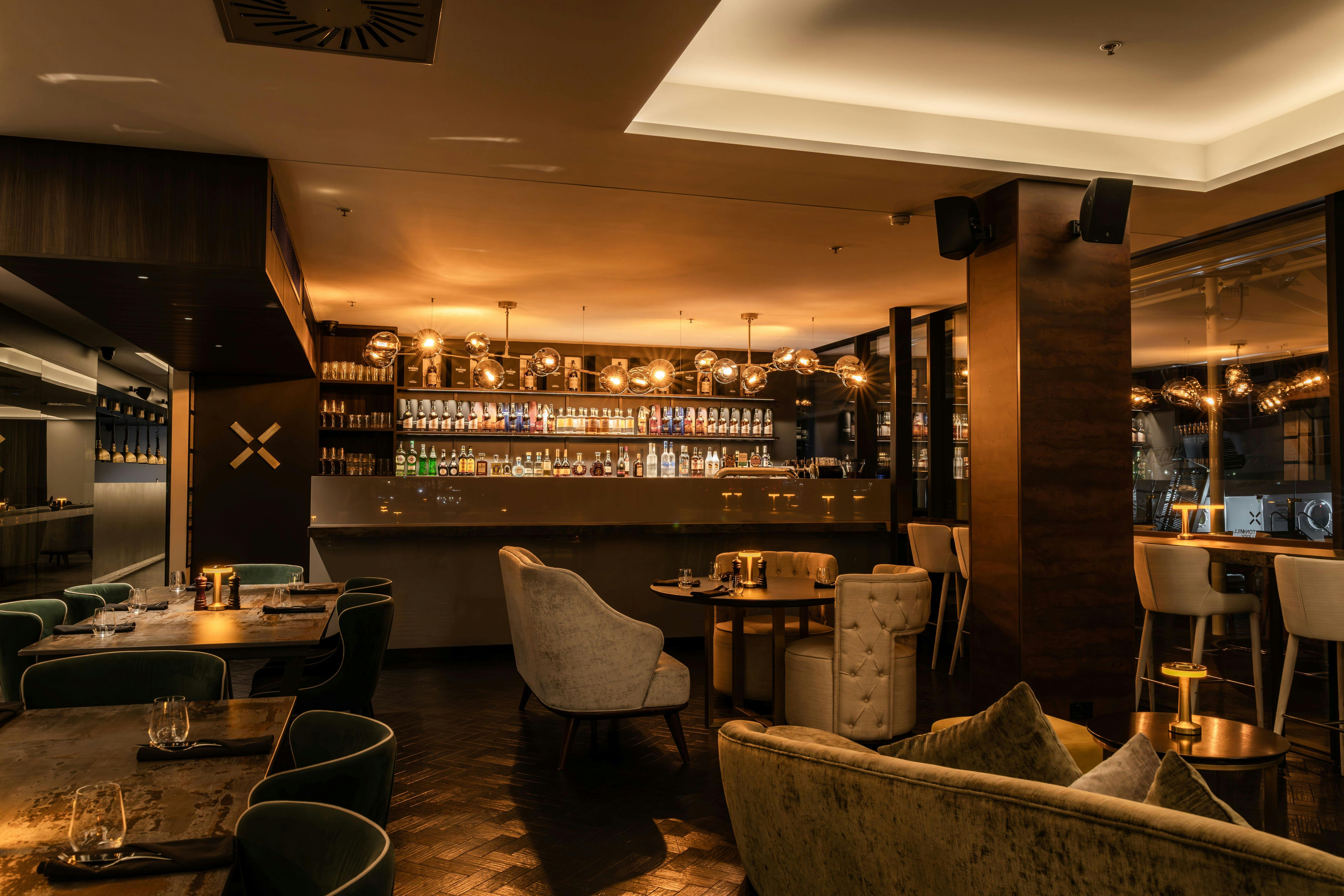A warmly lit bar with dining tables and arm chairs in the foreground