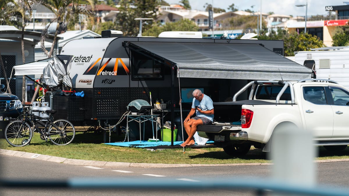 Image of a man sitting on his ute tray with caravan set up in background