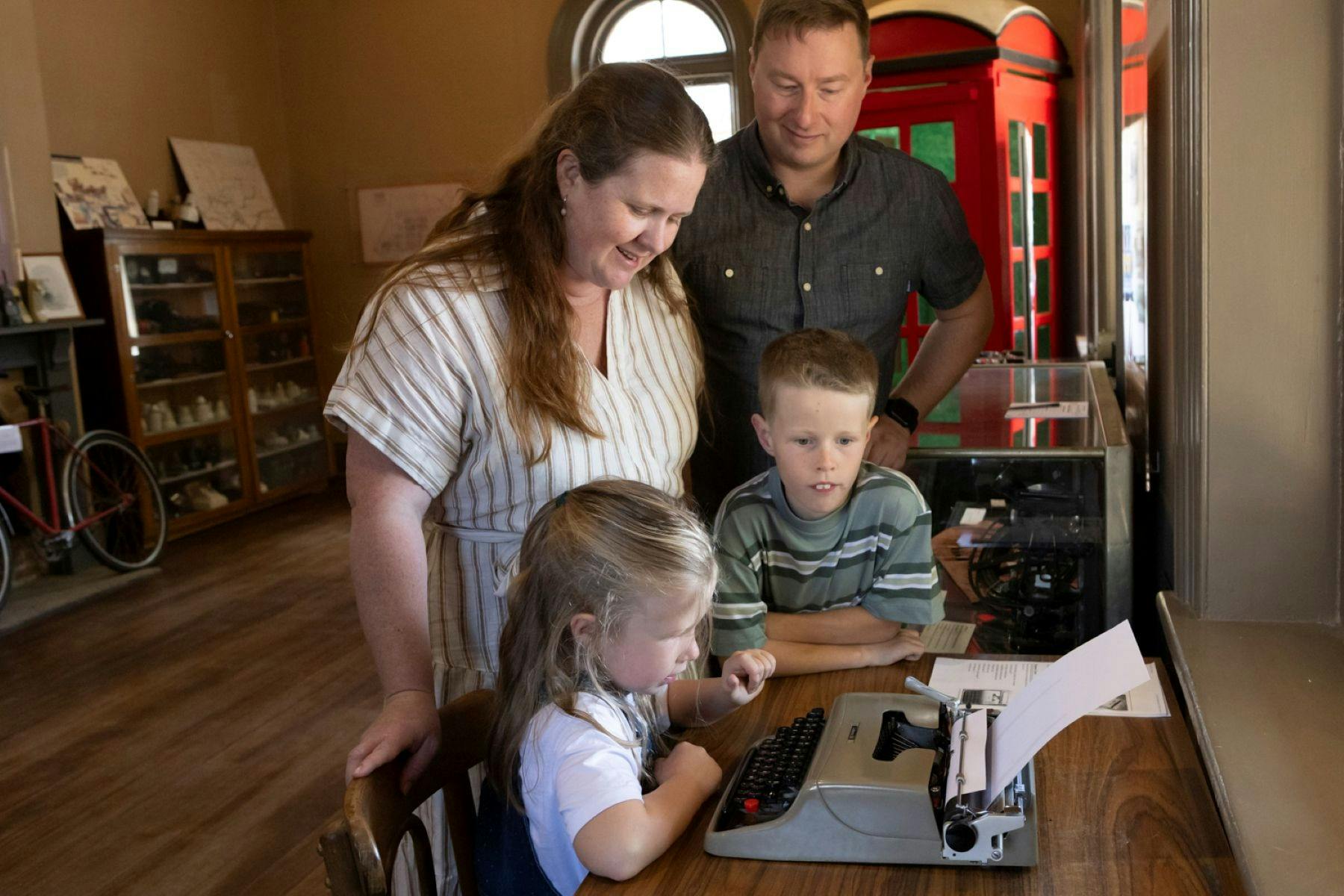 A mother, father, young girl and young boy crowd around an old telegraph machine