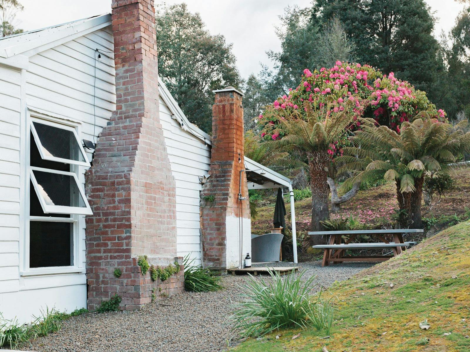 Outside of the cottage with brick chimneys, outdoor shower and stone bath tub adjacent to garden.