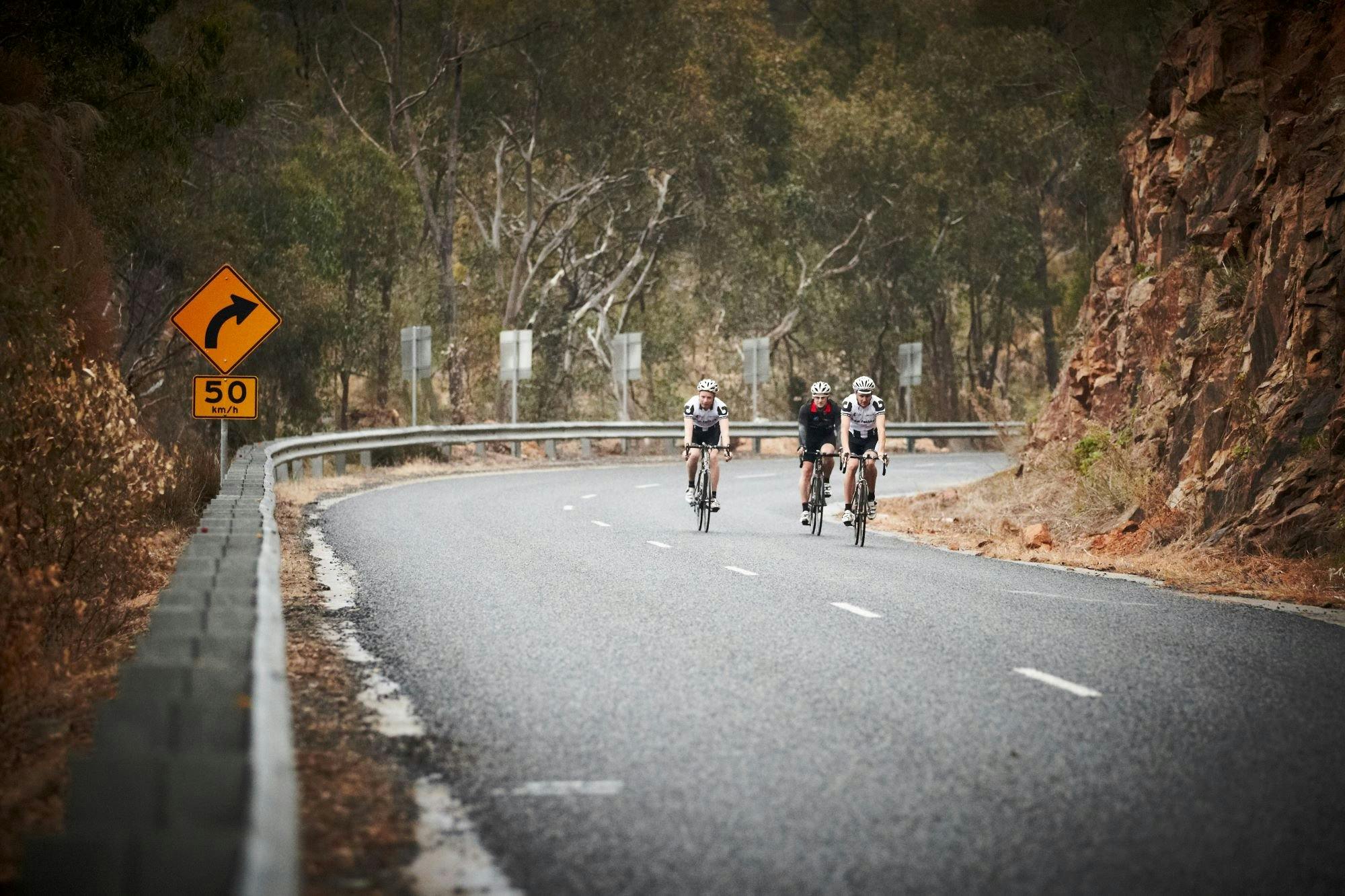 Cyclists road riding down a hill gum trees in background rocky cliff face on right hand side
