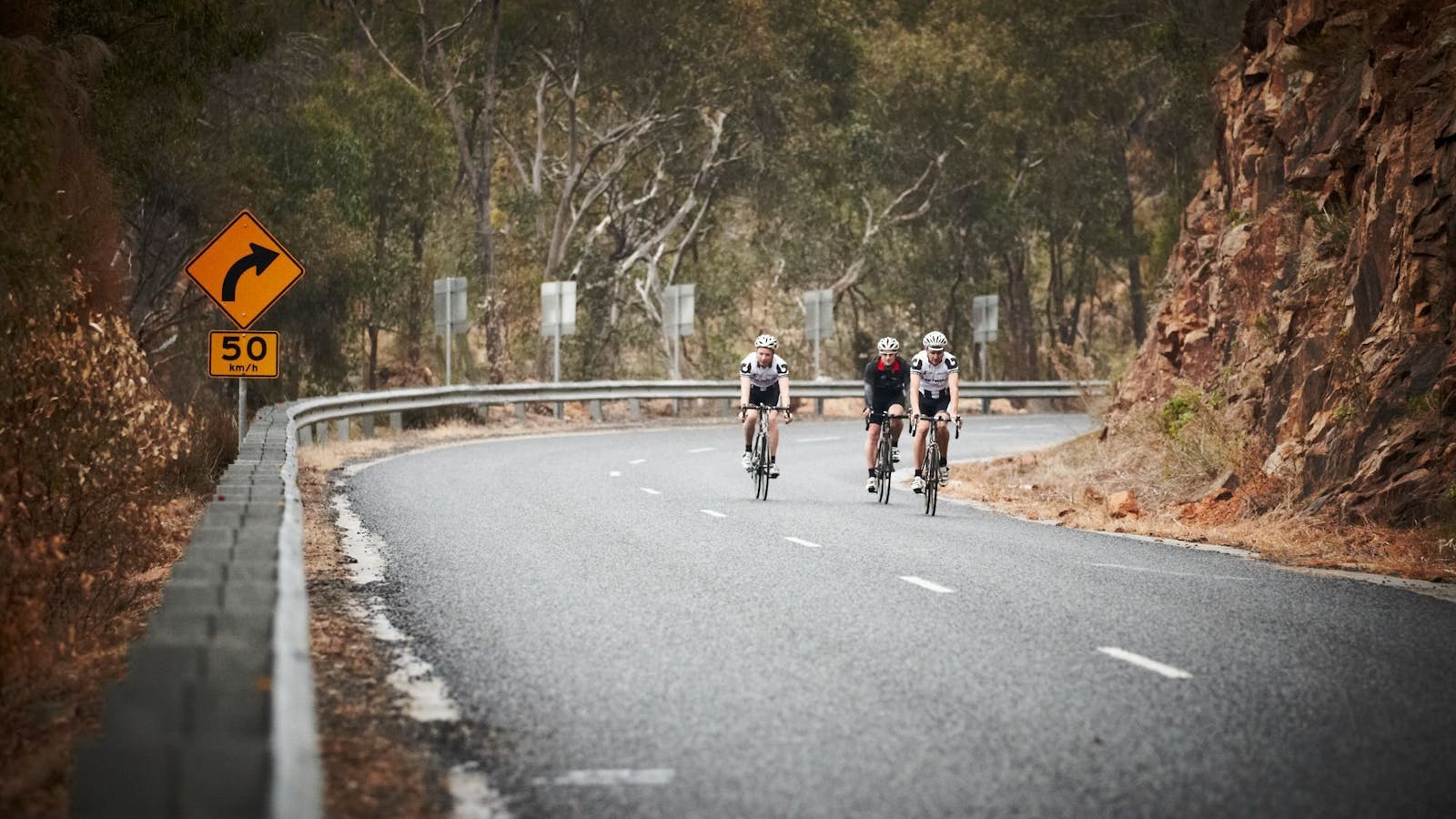Cyclists road riding down a hill gum trees in background rocky cliff face on right hand side
