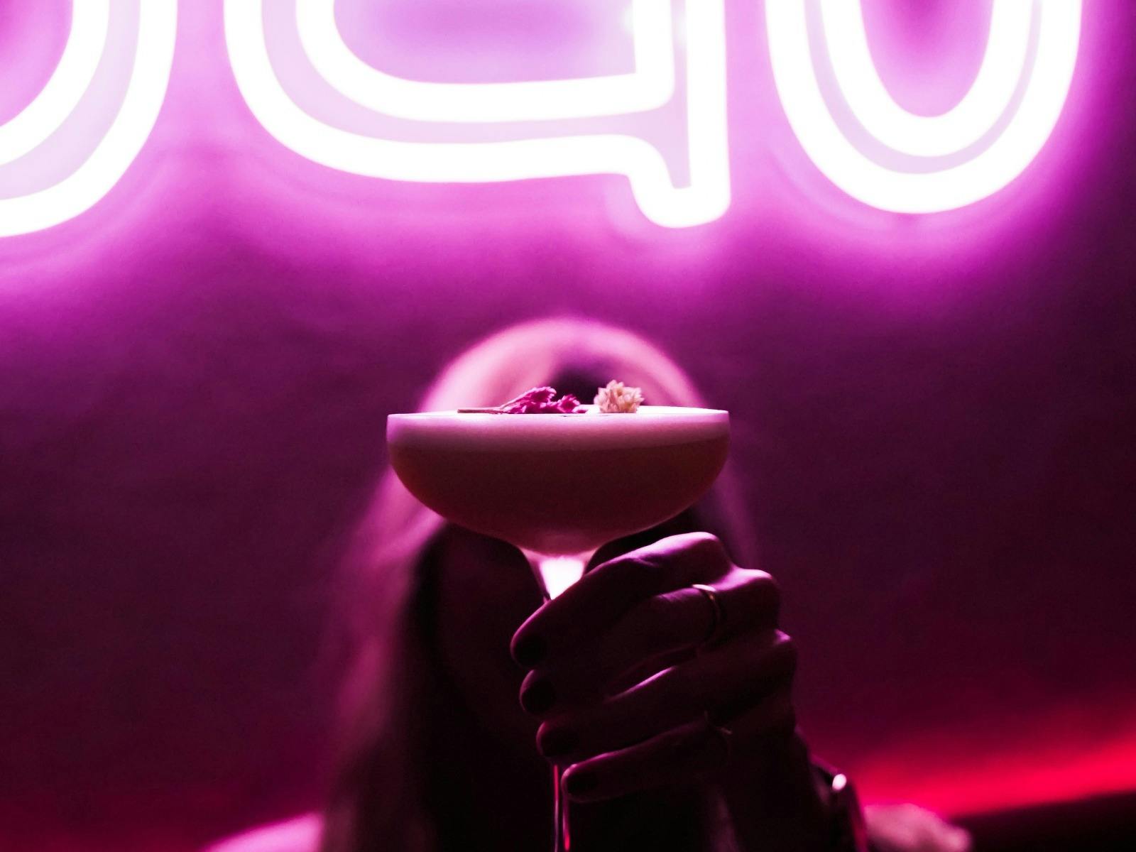 In front of the restaurant sign, a woman holds the signature cocktail. In front of the restaurant sign, a woman holds the signature cocktail.