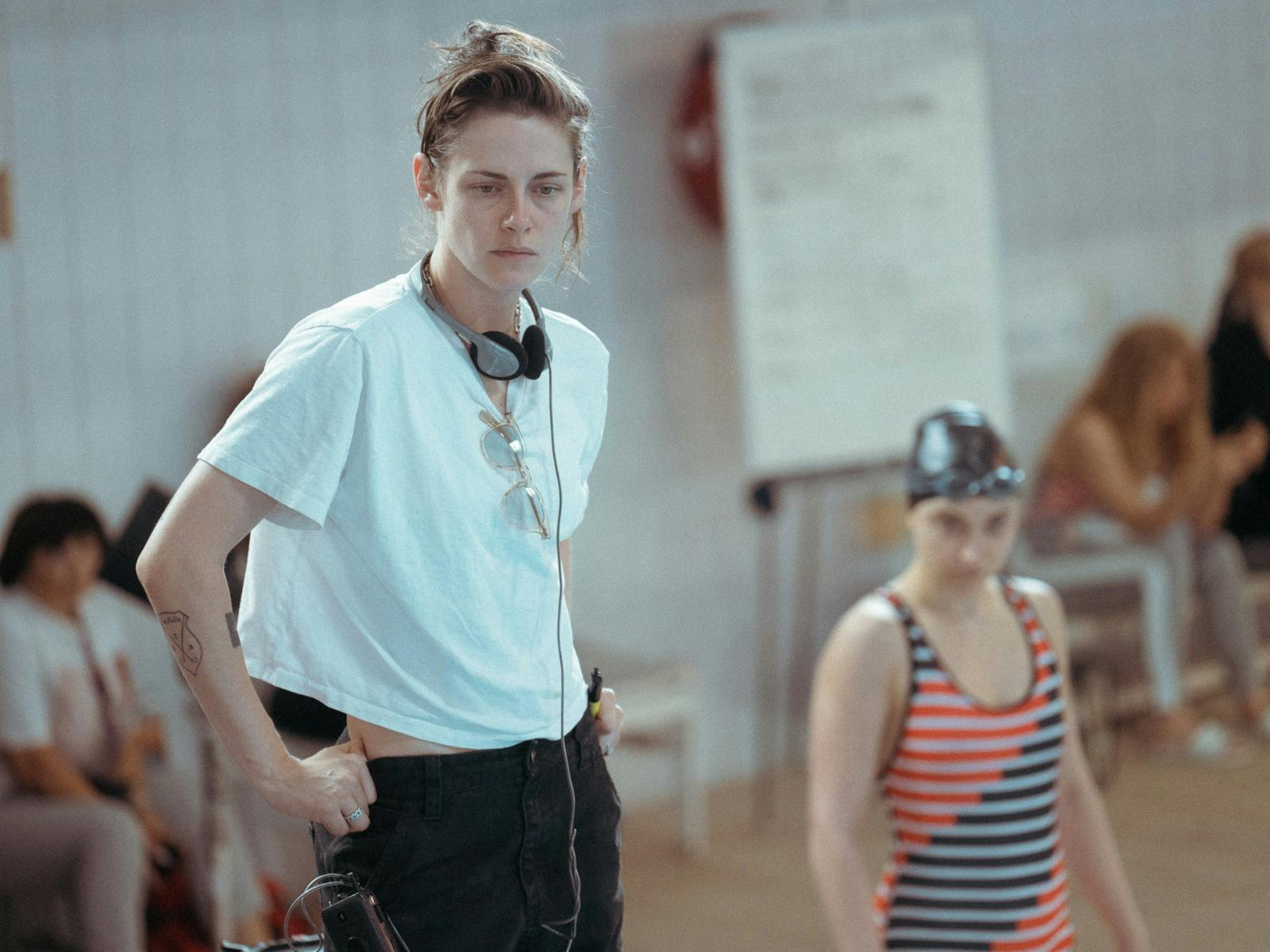 Woman stands with headphones and white t shirt with hands in pockets looking forlorn