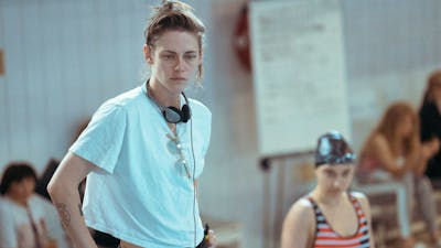 Woman stands with headphones and white t shirt with hands in pockets looking forlorn
