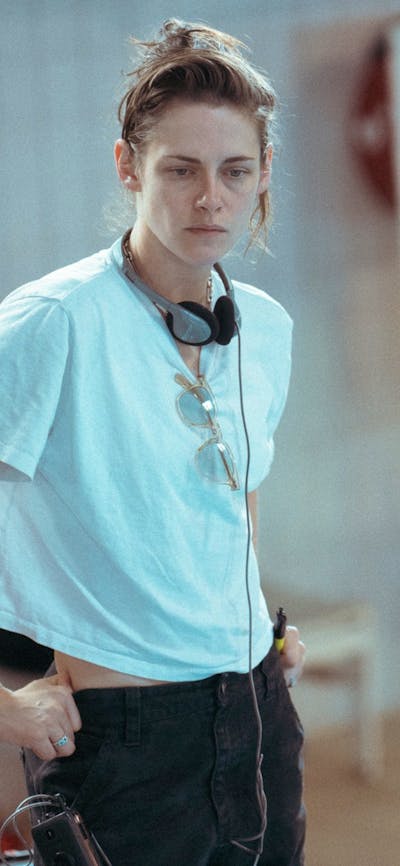 Woman stands with headphones and white t shirt with hands in pockets looking forlorn