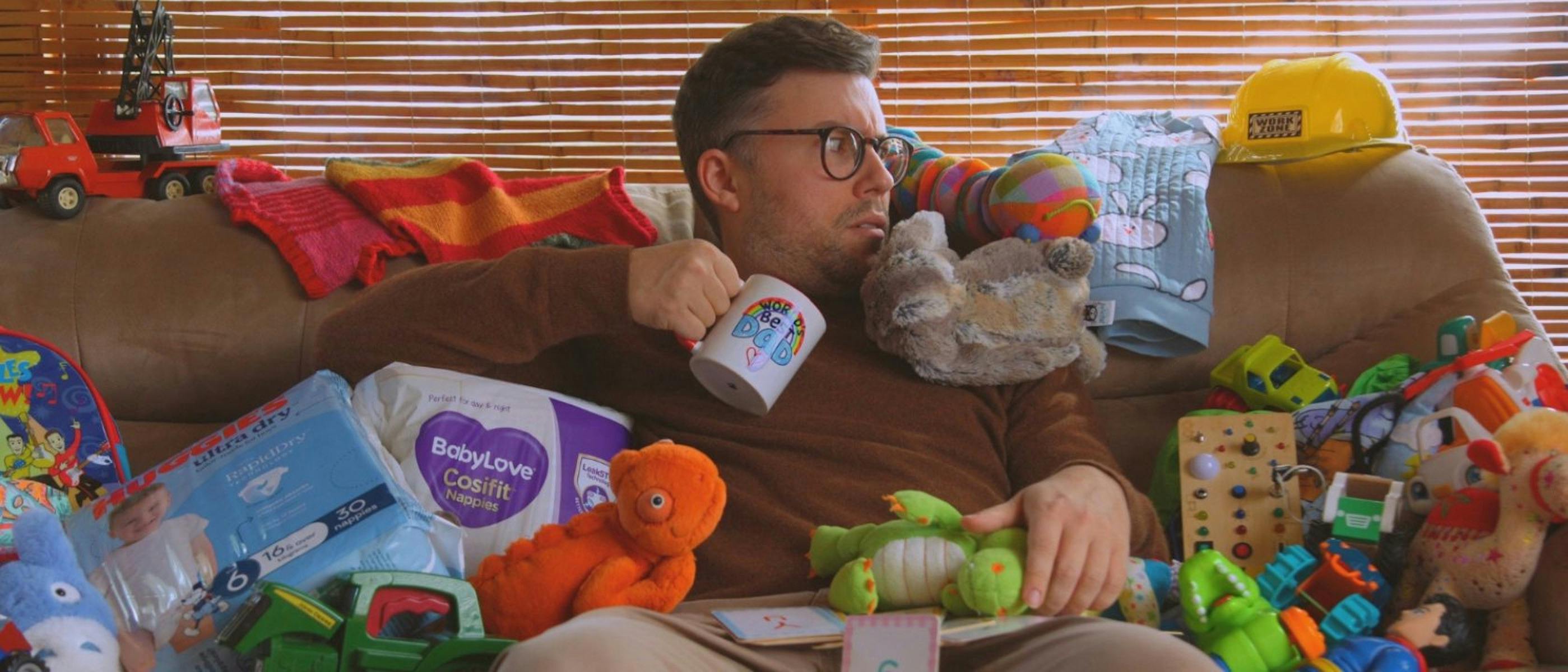 A man holding a coffee cup sits on a couch, covered in stuffed toys and packets of nappies