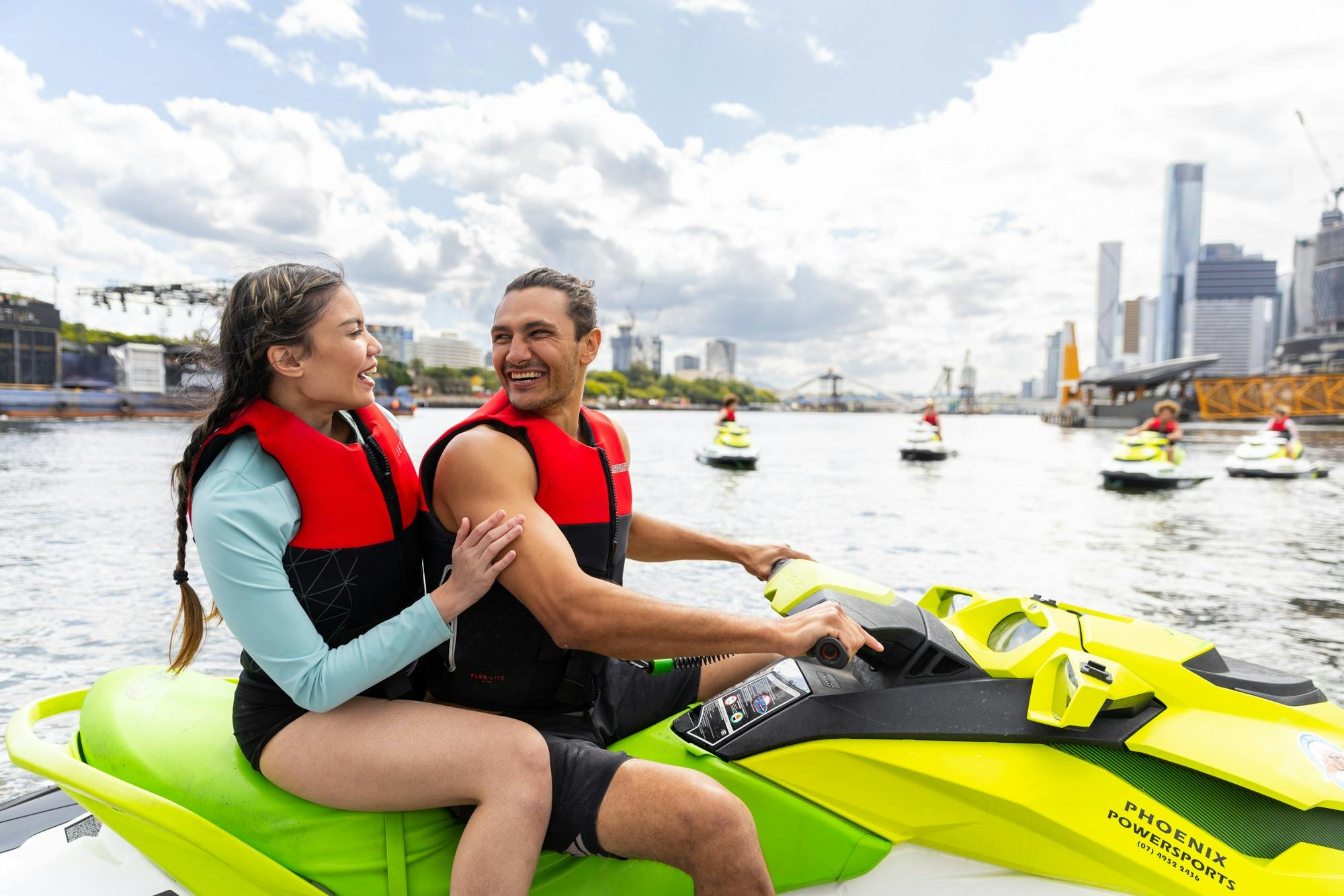 A couple on a jet ski smiling at each other on the Brisbane River with Brisbane City in background.