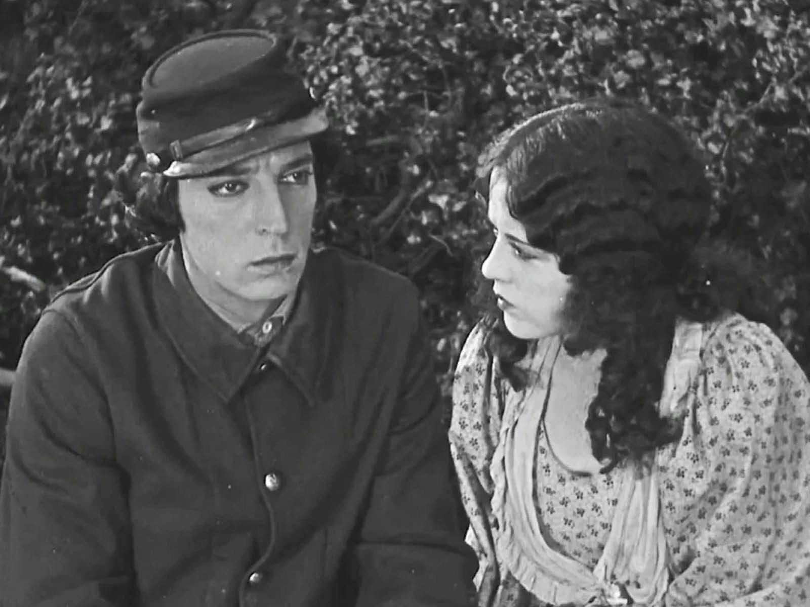 Buster Keaton and his love interest sit together outside on a bench with a leafy green bush directly