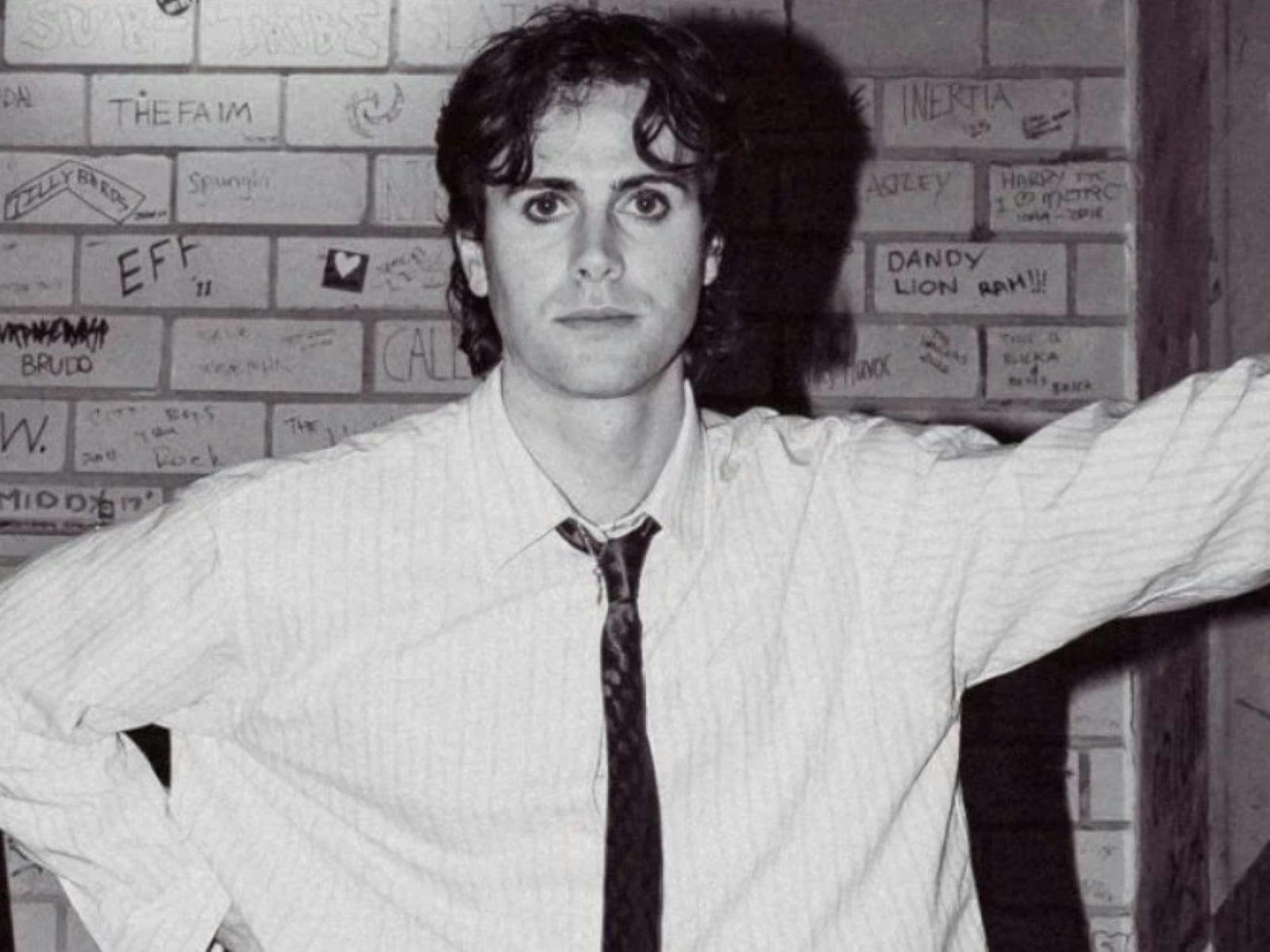 Yes Boon in a suit and tie leans up against a stairwell