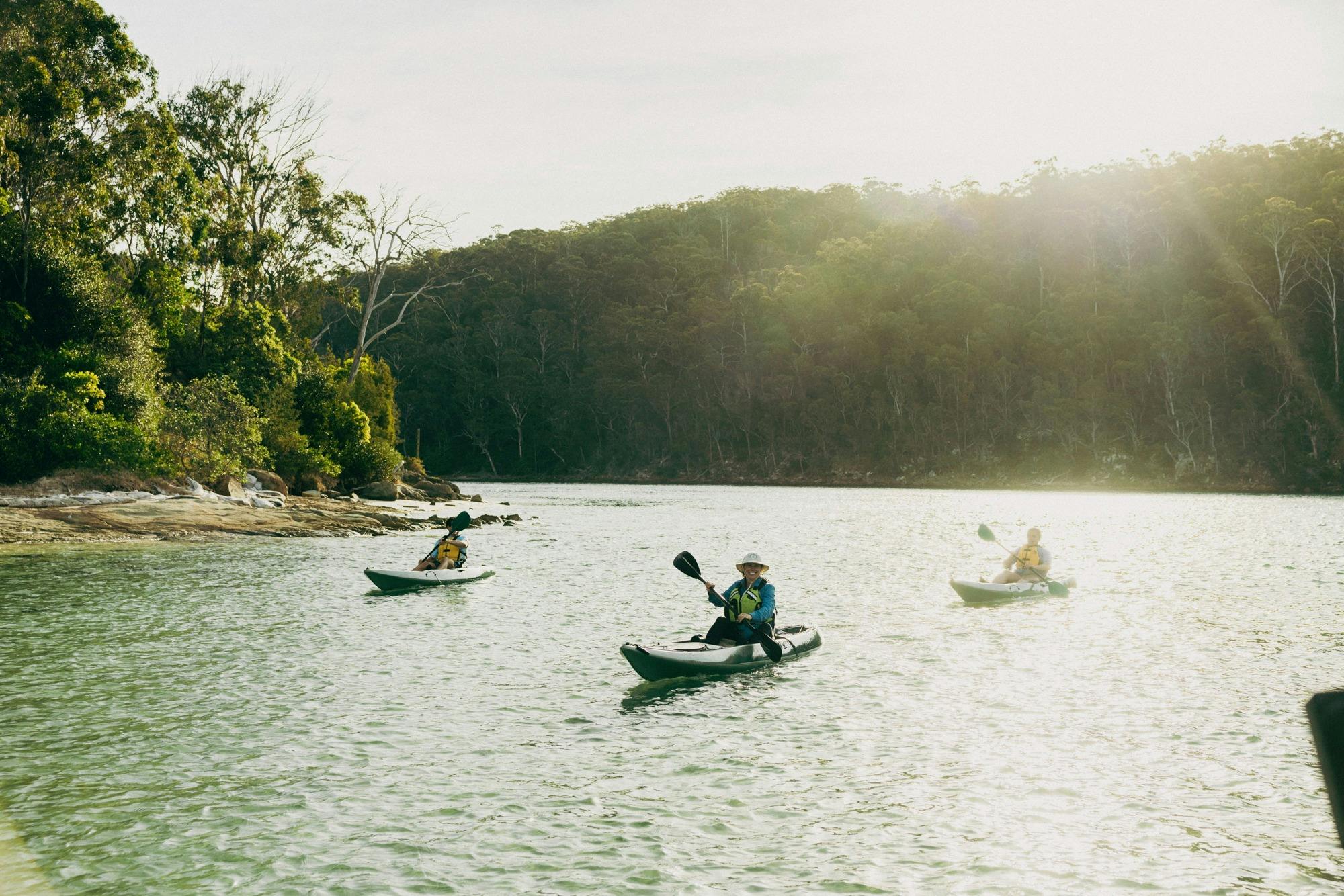 Three guests in kayaks paddling along the calm Pambula River, illuminated by warm, golden sunset lig