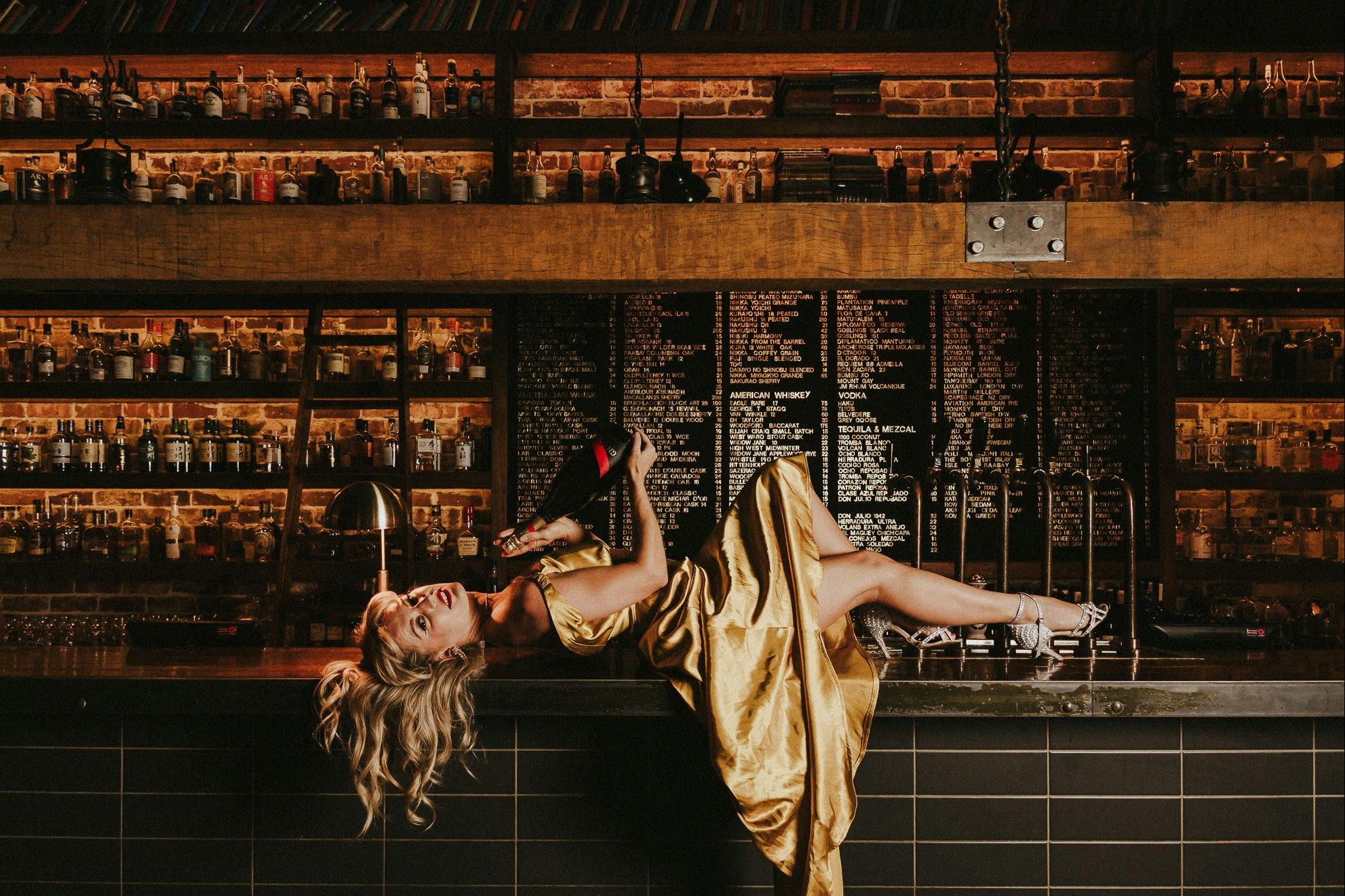 A girl in a golden dress, lays atop a bar holding champagne