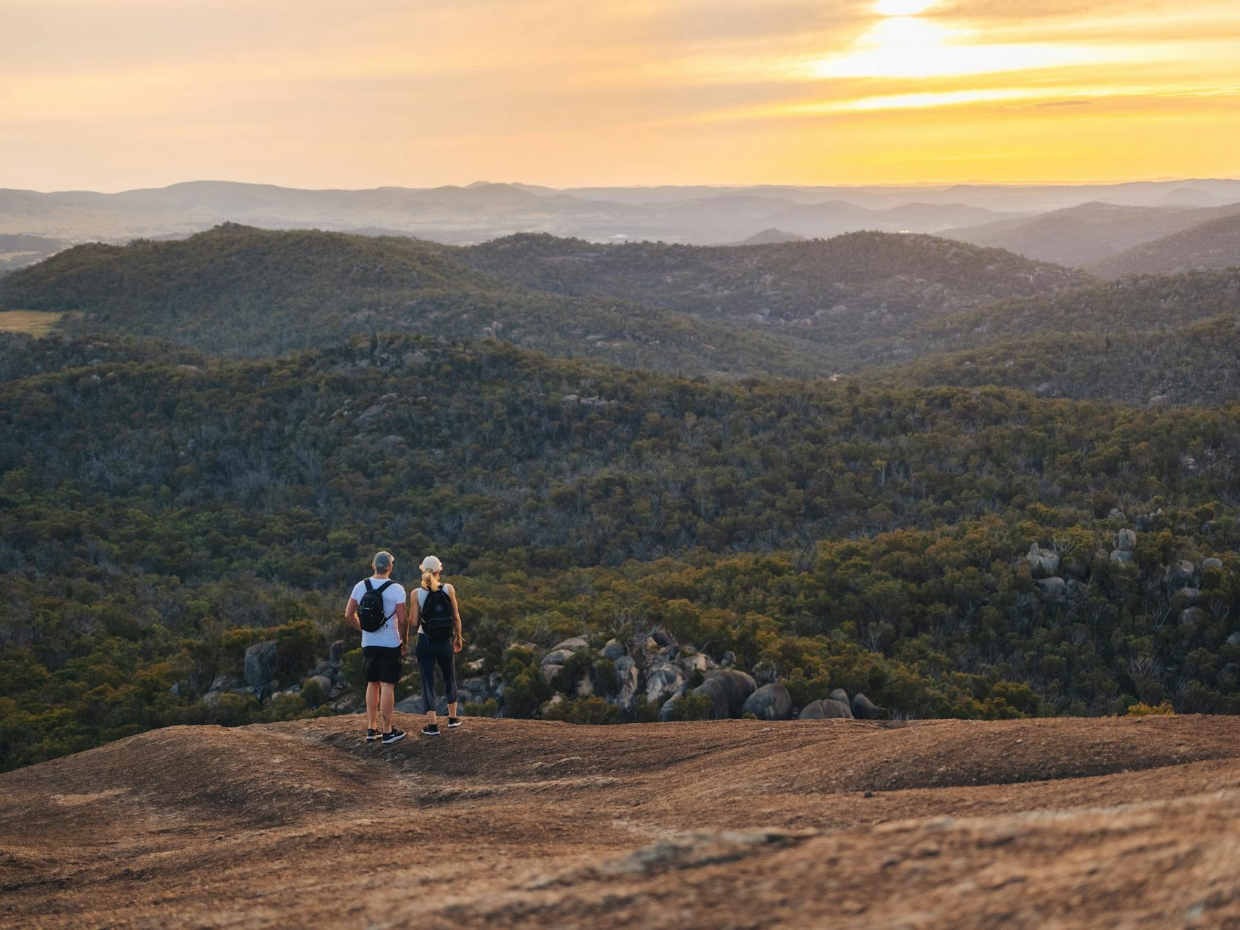 Two hikers standing on edge of granite mountain looking out into the distance and sunset.