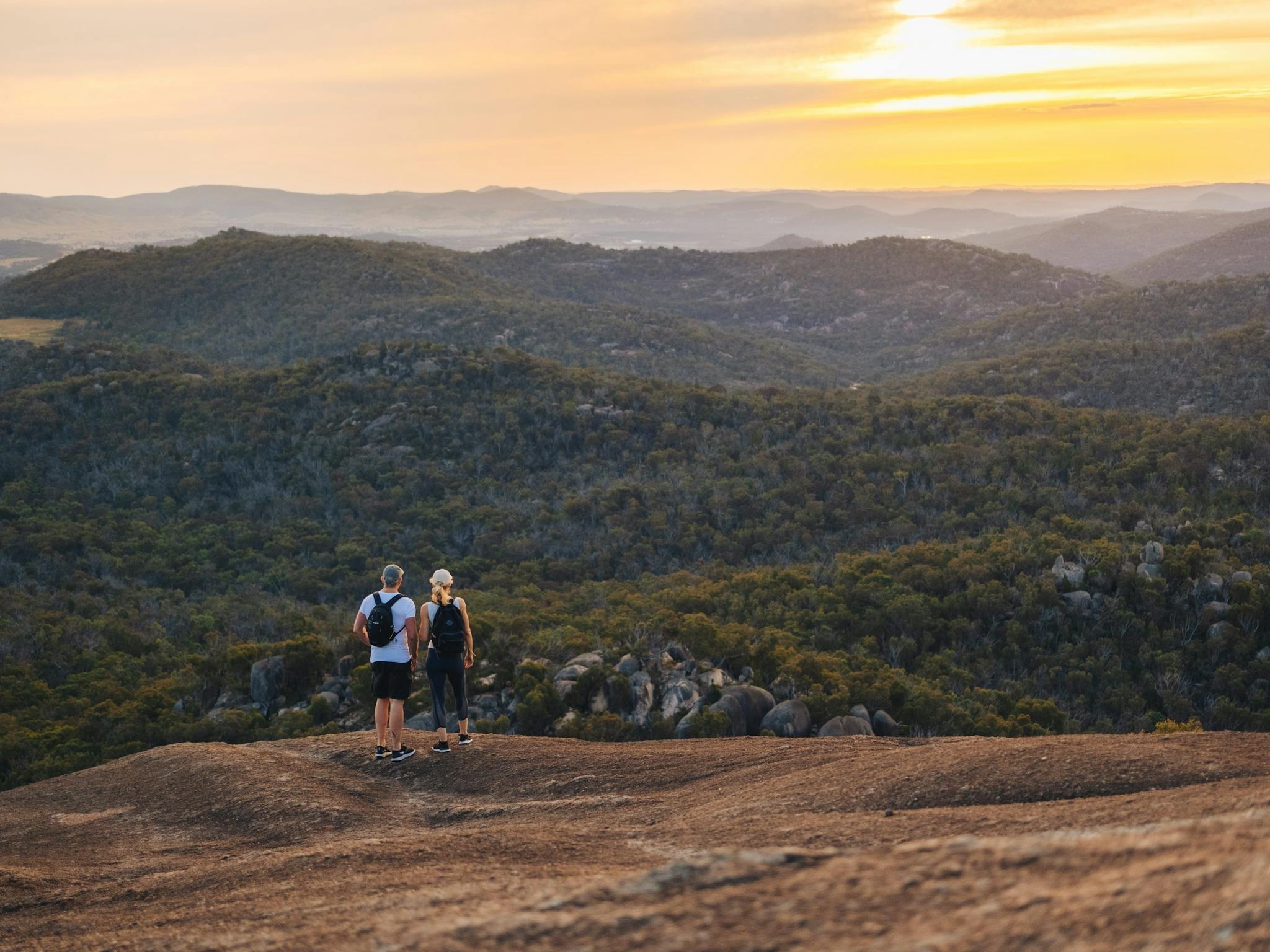 Two hikers standing on edge of granite mountain looking out into the distance and sunset.