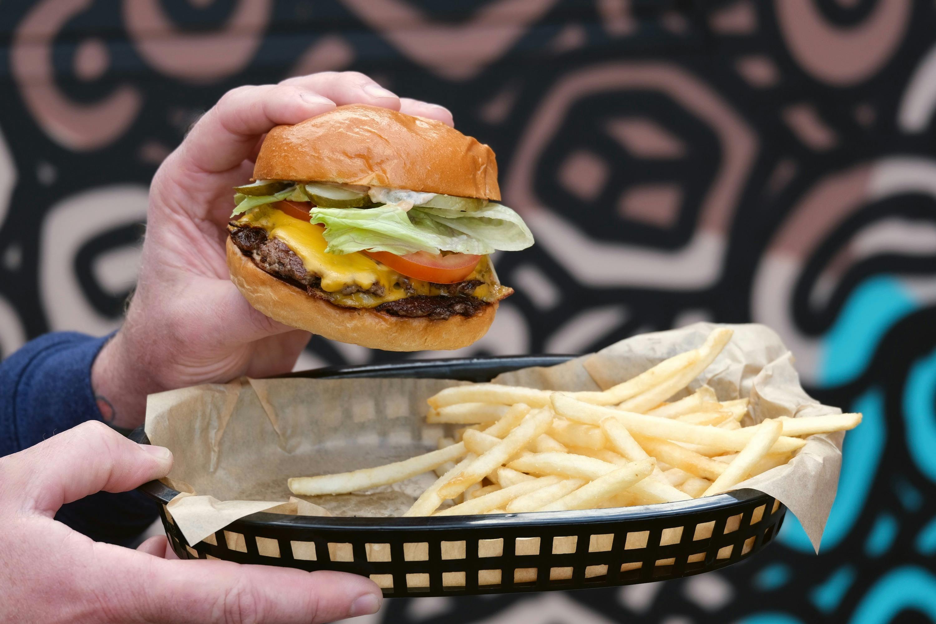 Man's hands holding  double cheeseburger with lettuce and tomato and  black plastic basket of fries