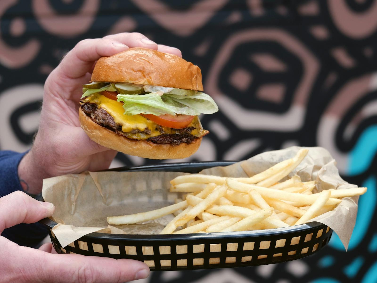 Man's hands holding  double cheeseburger with lettuce and tomato and  black plastic basket of fries