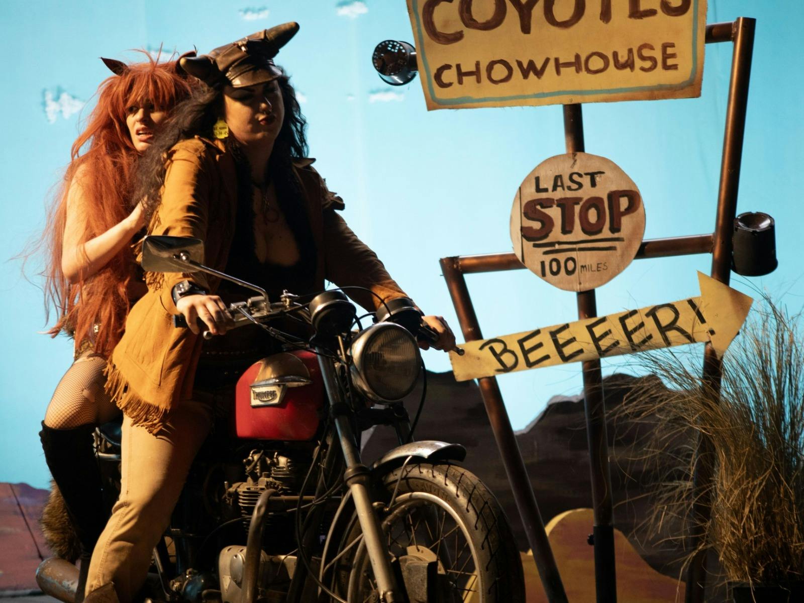 Two women dressed in costumes and wigs on a motorcycle in a theatre like setting